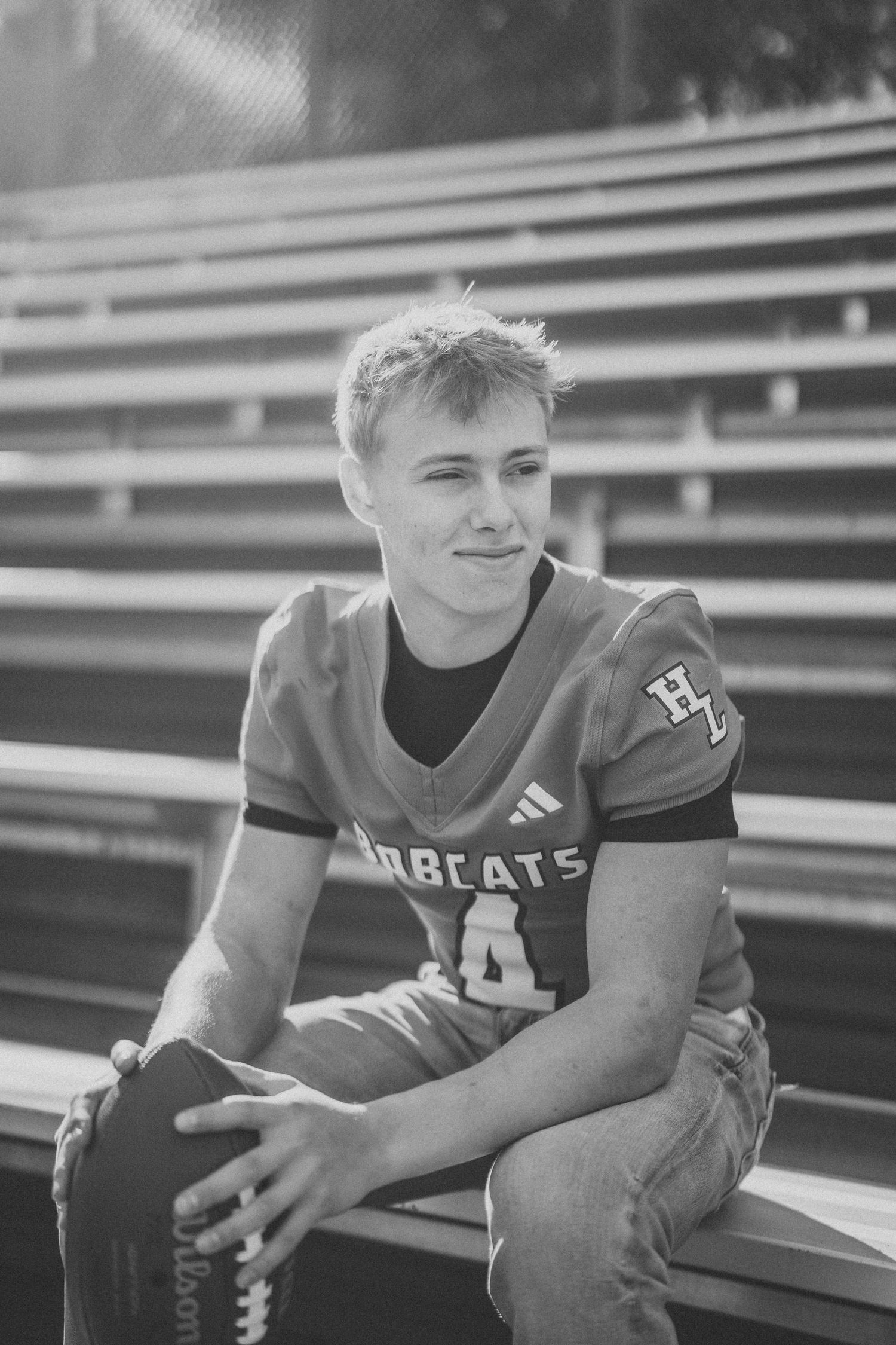 Black and white photo of football player sitting on bleachers in uniform.