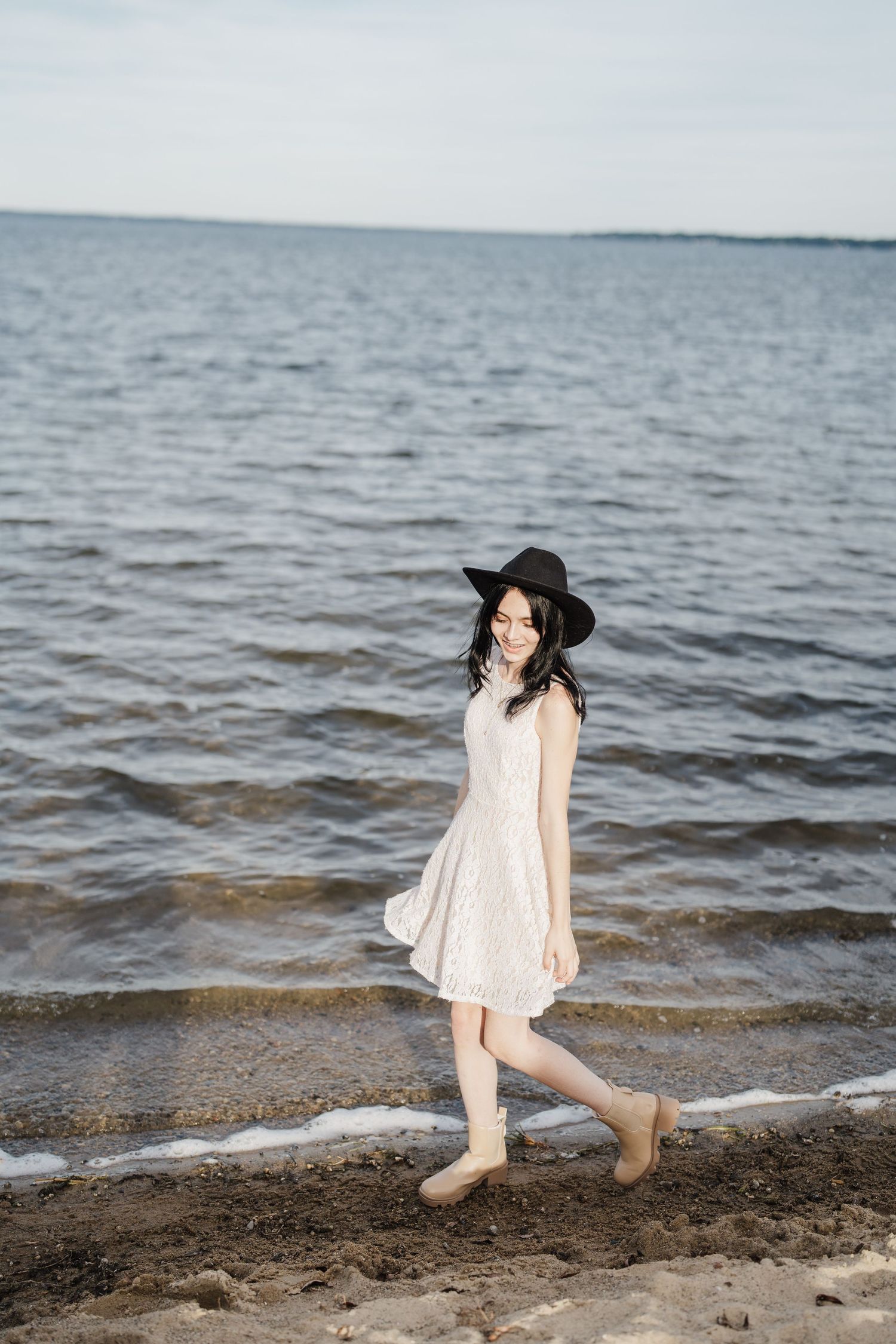 A person in a flowing white dress and black hat walks playfully along a rocky beach shoreline at dusk.