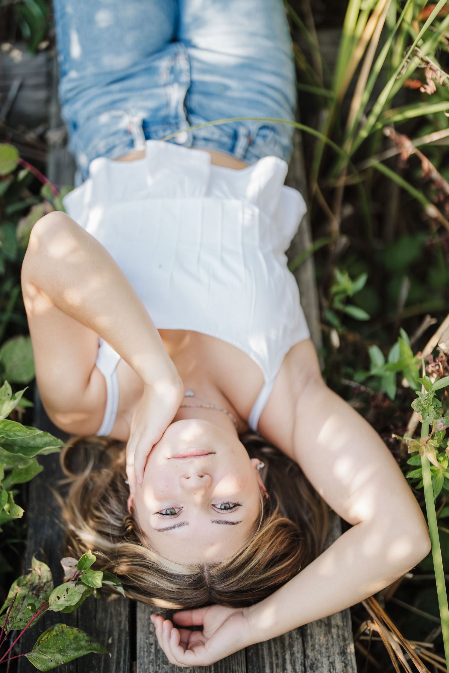 Person lying on wooden surface outdoors wearing white top and denim jeans in natural lighting.