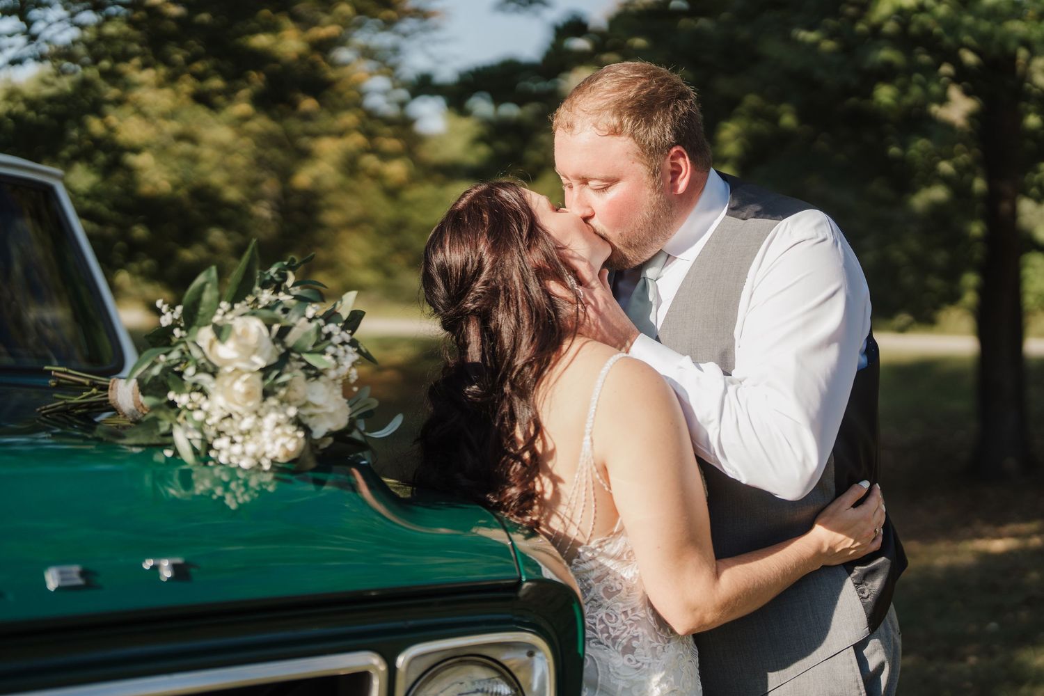 A couple shares a romantic kiss next to a vintage green truck with a white floral bouquet on a sunny day.
