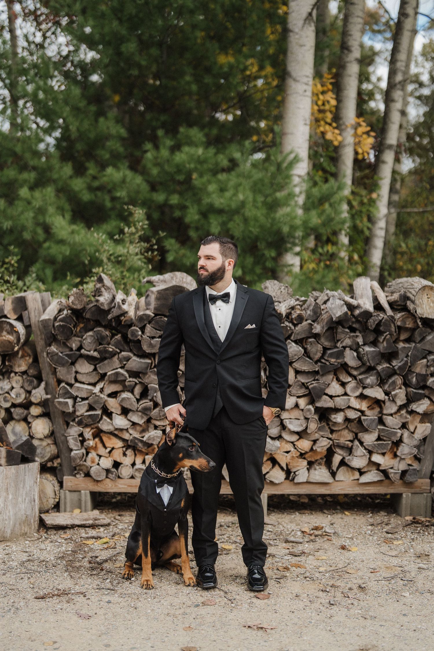 A groom in a black tuxedo sits on a wooden bench with a dog beside stacks of firewood in a rustic outdoor setting.