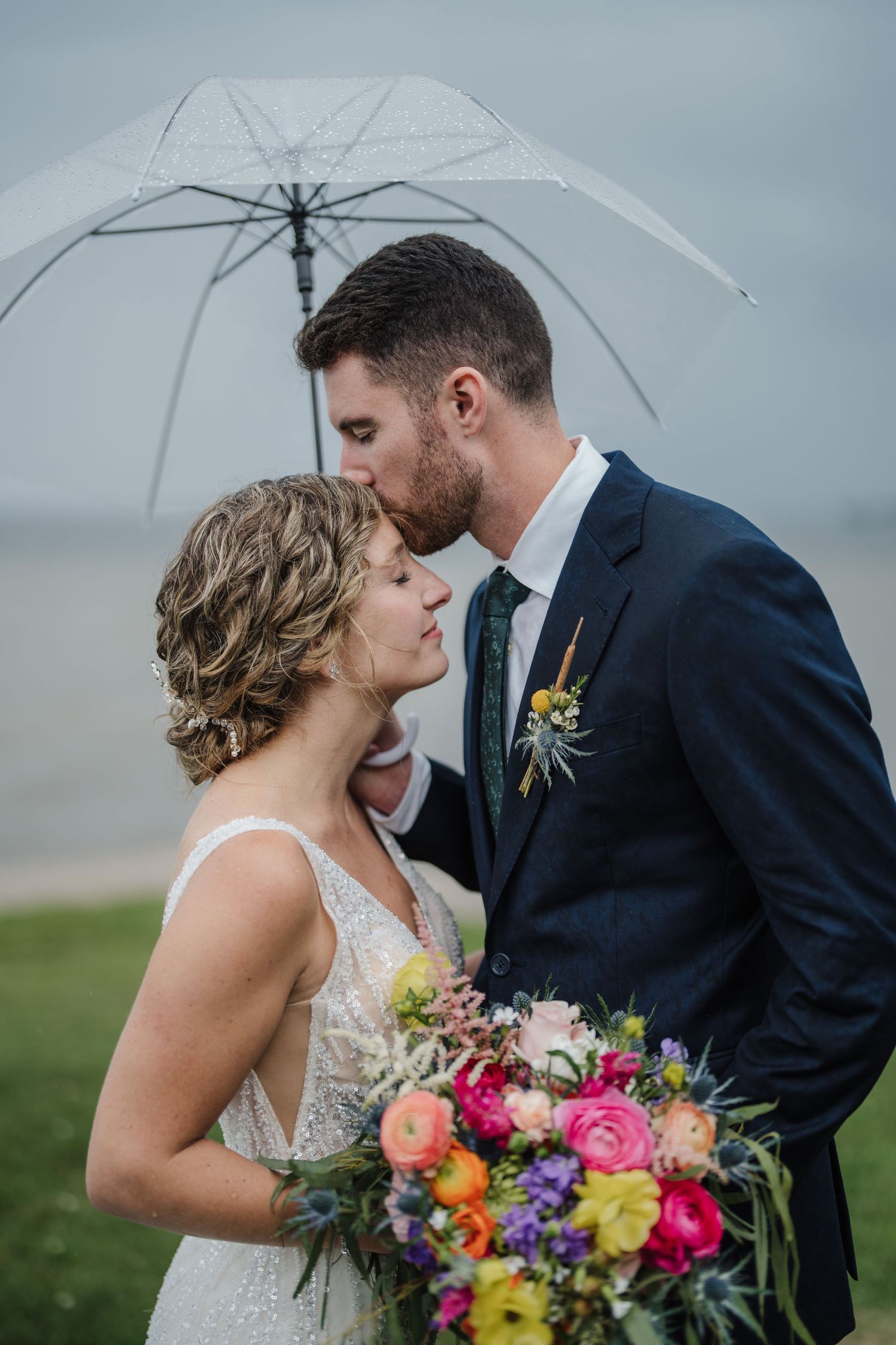 A couple embraces under a clear umbrella while holding a vibrant bouquet against a cloudy backdrop.