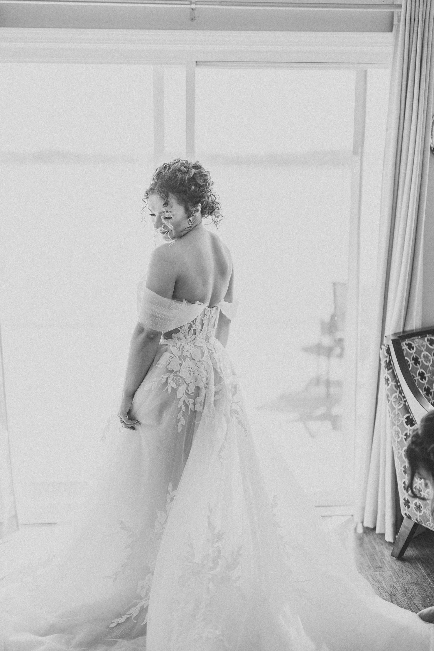 Black and white photo of a person in a lace wedding dress looking over their shoulder by a window.