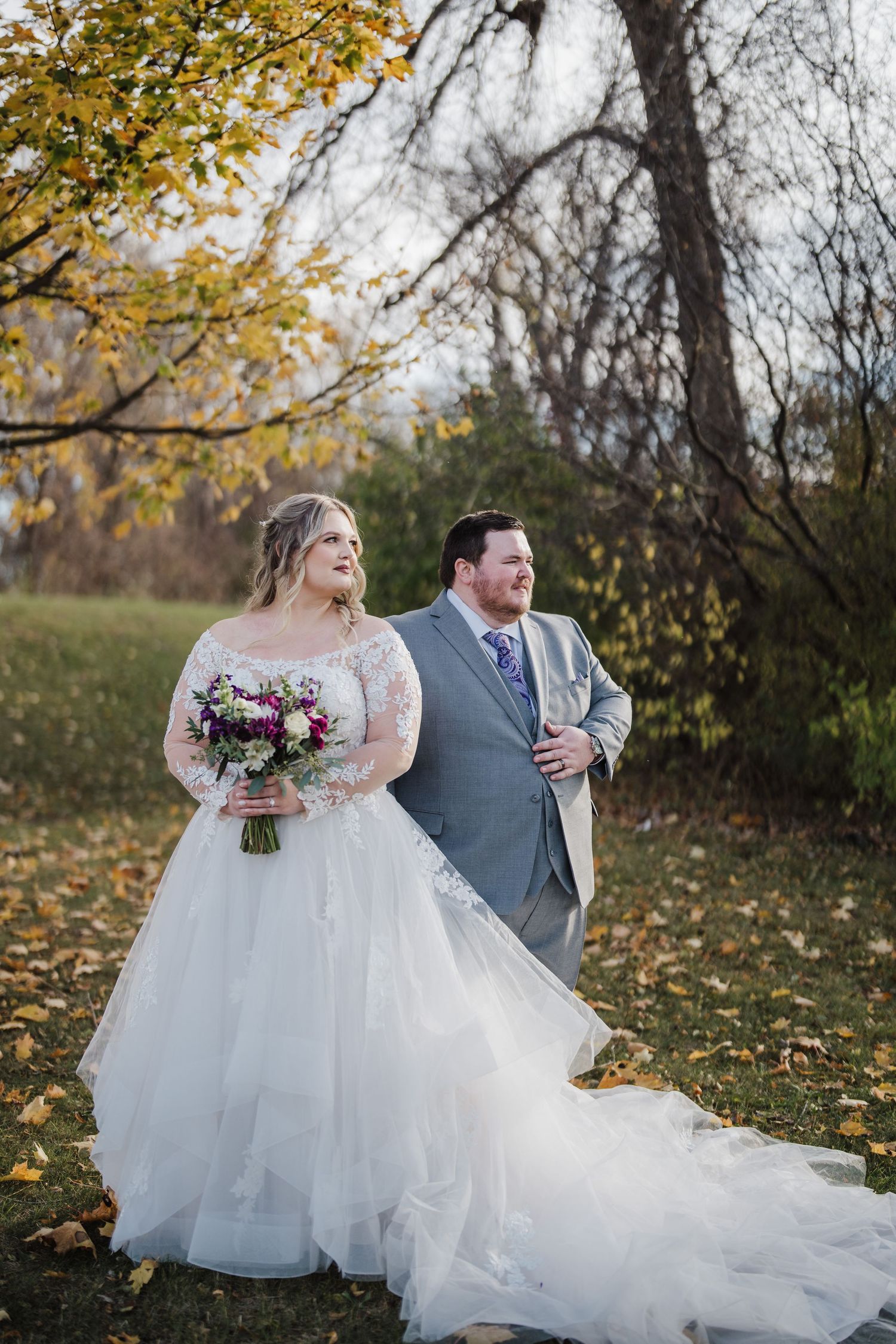 Wedding couple poses outdoors with fall foliage backdrop and purple bouquet.