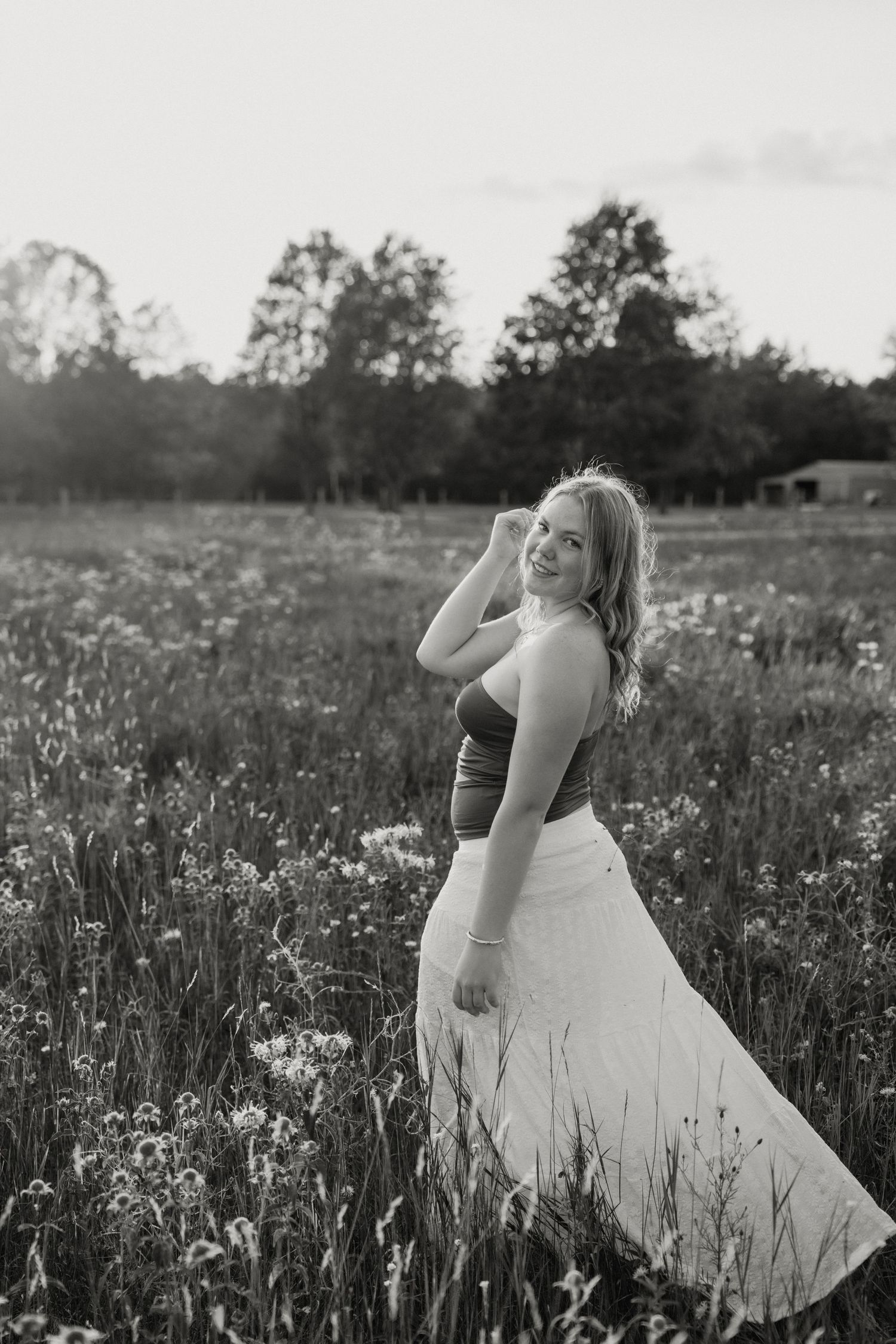 Black and white photo of a smiling young woman in a flowy skirt standing in a wildflower field at golden hour.