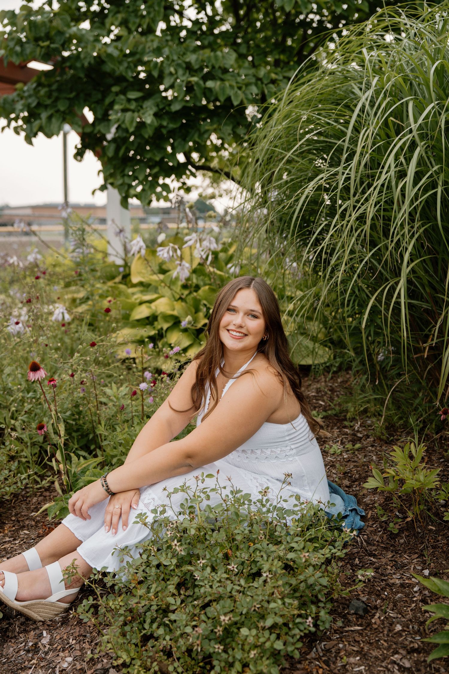 Young woman in white dress sitting among garden flowers and tall grasses, smiling outdoors.