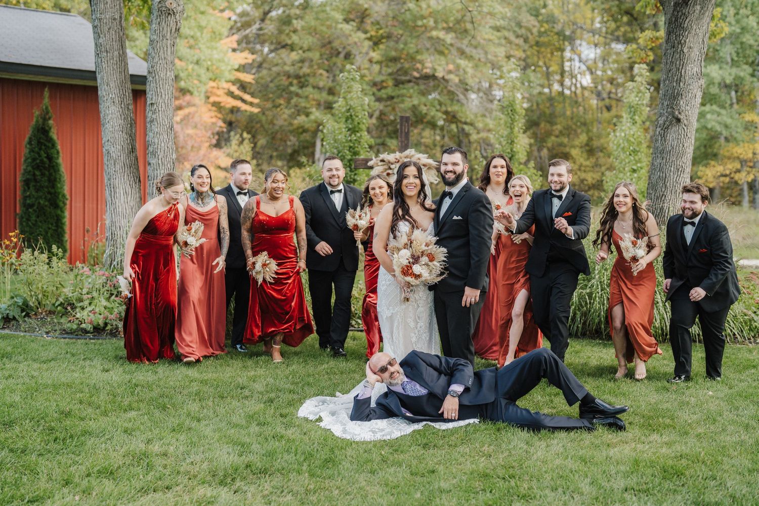 A wedding party poses in front of a rustic red barn and autumn trees in coordinating burgundy attire.