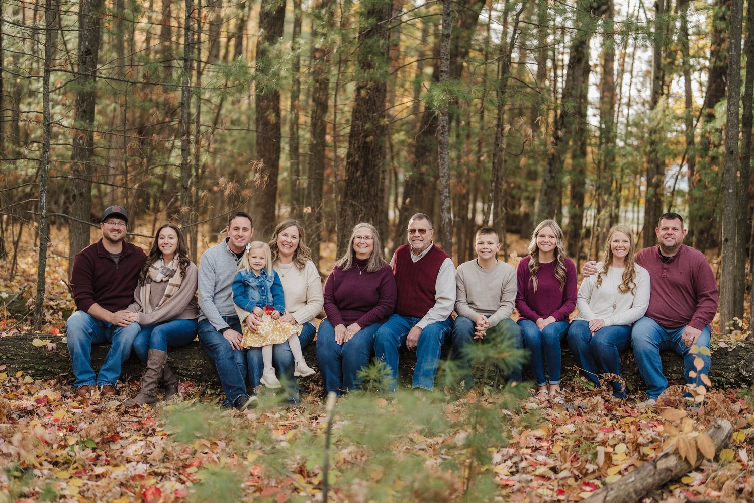 A group sits together on fallen autumn leaves in a wooded area for outdoor family photos.