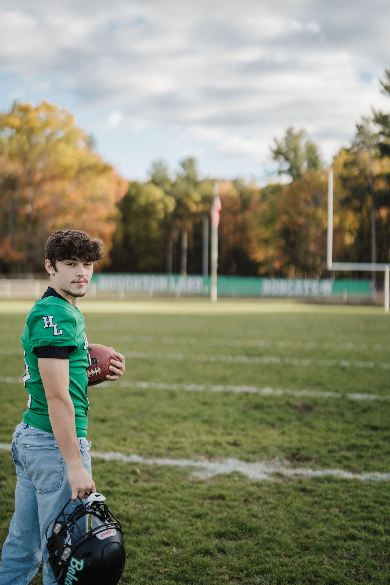 Player in green Bobcats uniform holds football and helmet on grassy sports field during fall season.