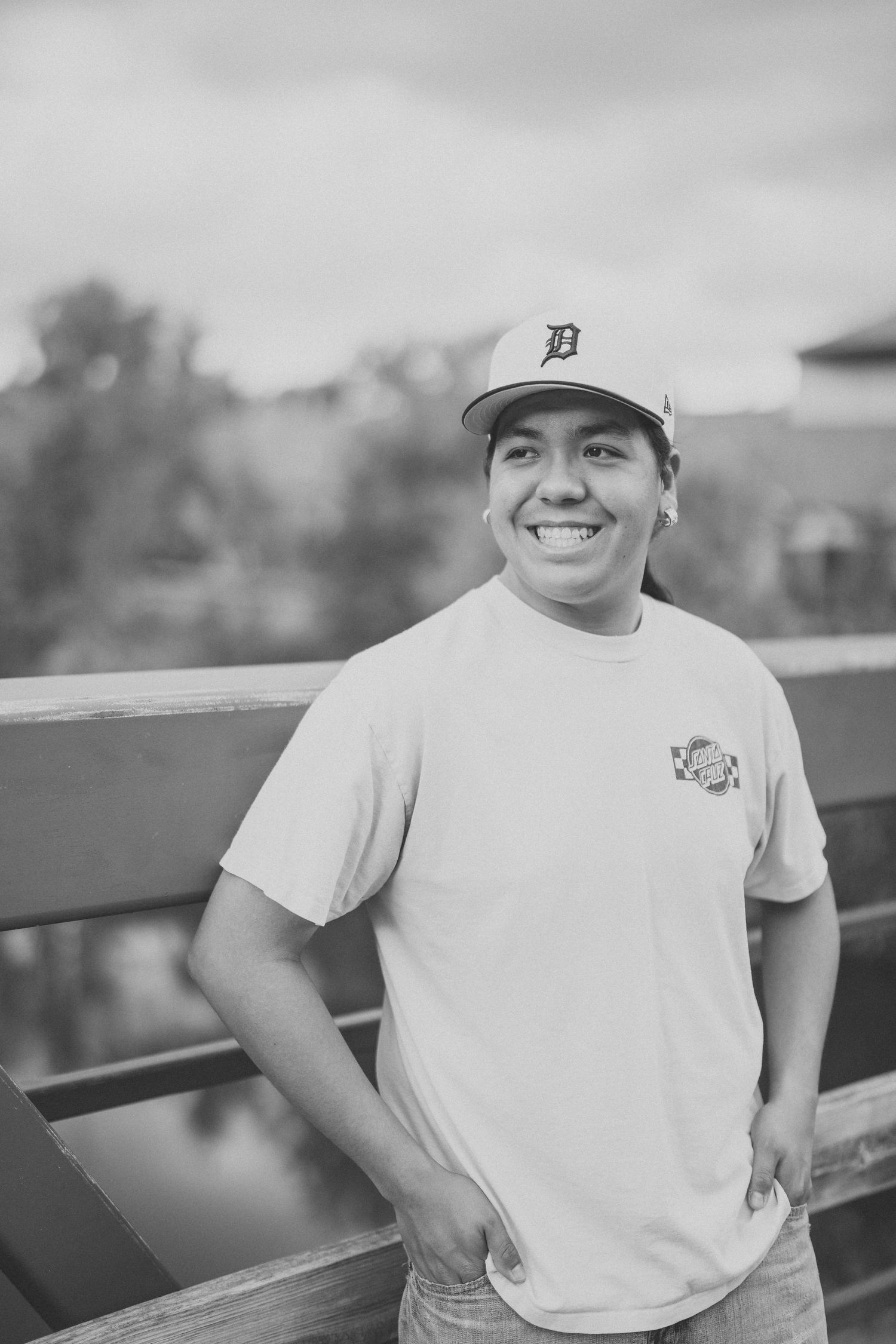 A black and white portrait of someone wearing a baseball cap and white t-shirt while leaning against a railing.