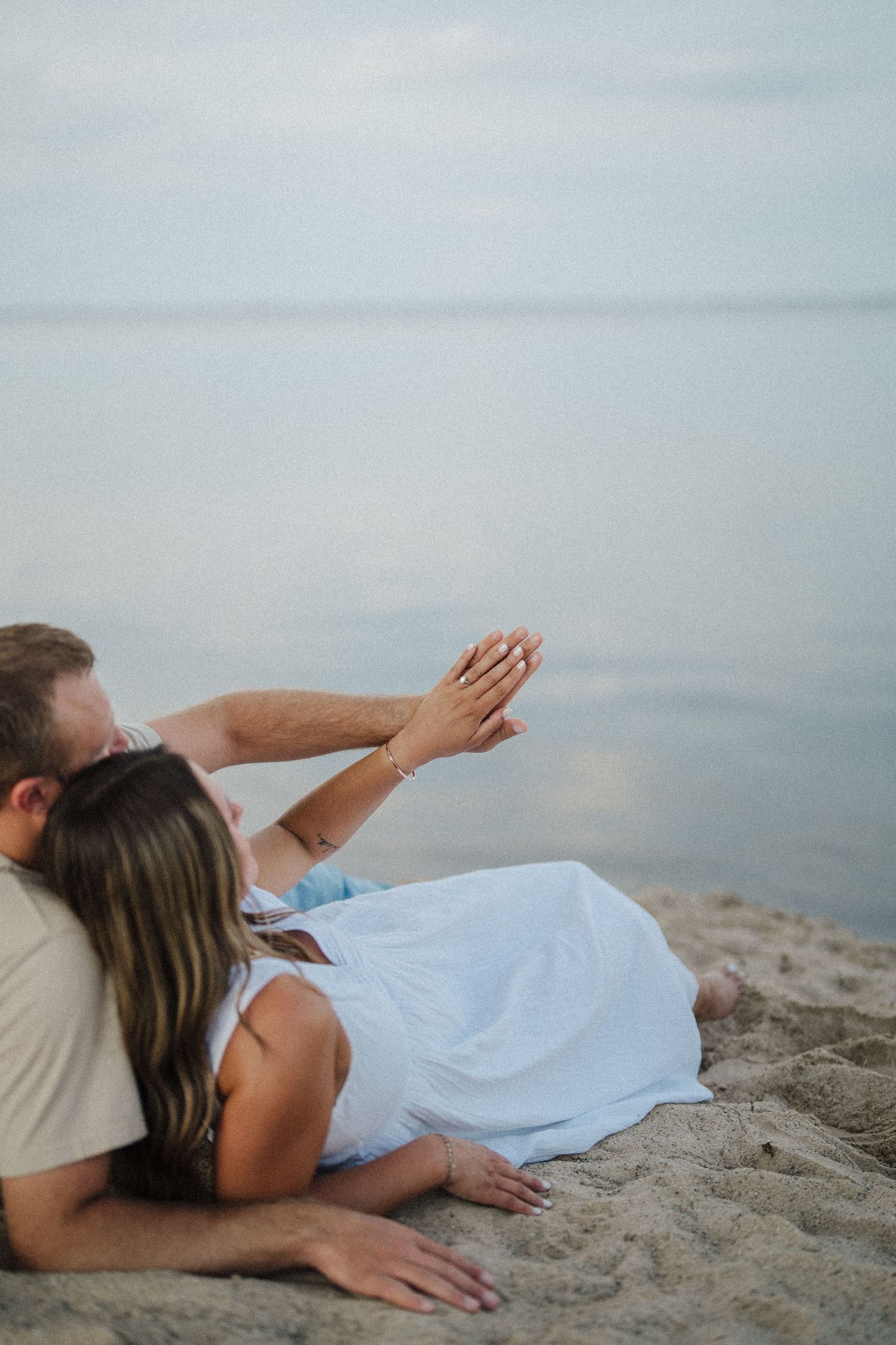 Couple lying on sandy beach at dusk reaching towards calm ocean waters.