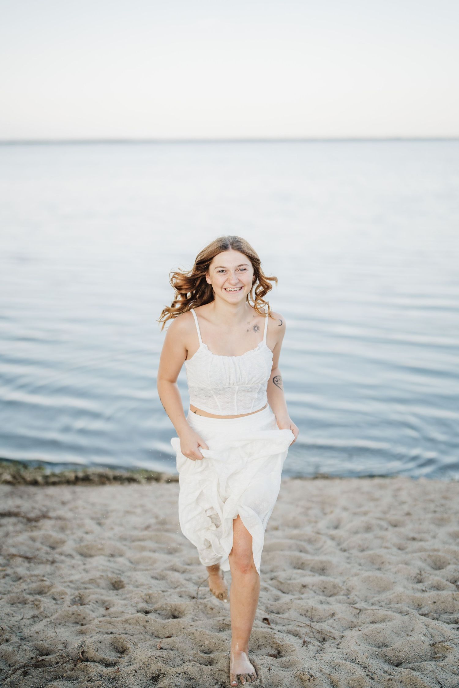 A person in a flowing white dress runs joyfully along a sandy beach near the ocean.