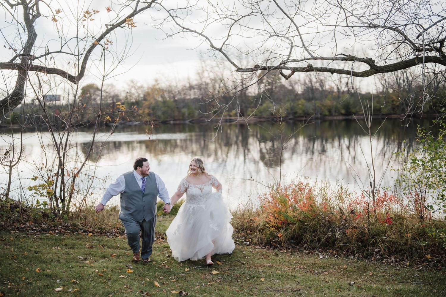 Couple walks hand in hand along lakeside path in wedding attire.
