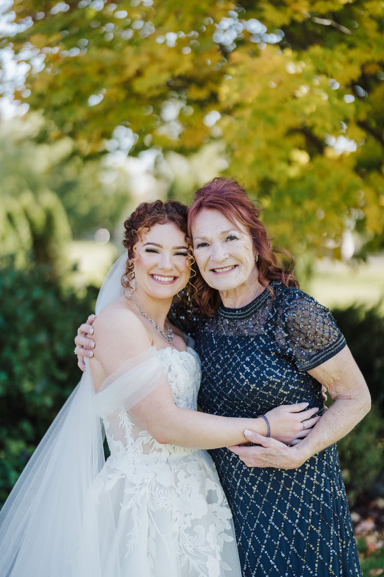 Two people embrace and smile outdoors under autumn trees with golden foliage.