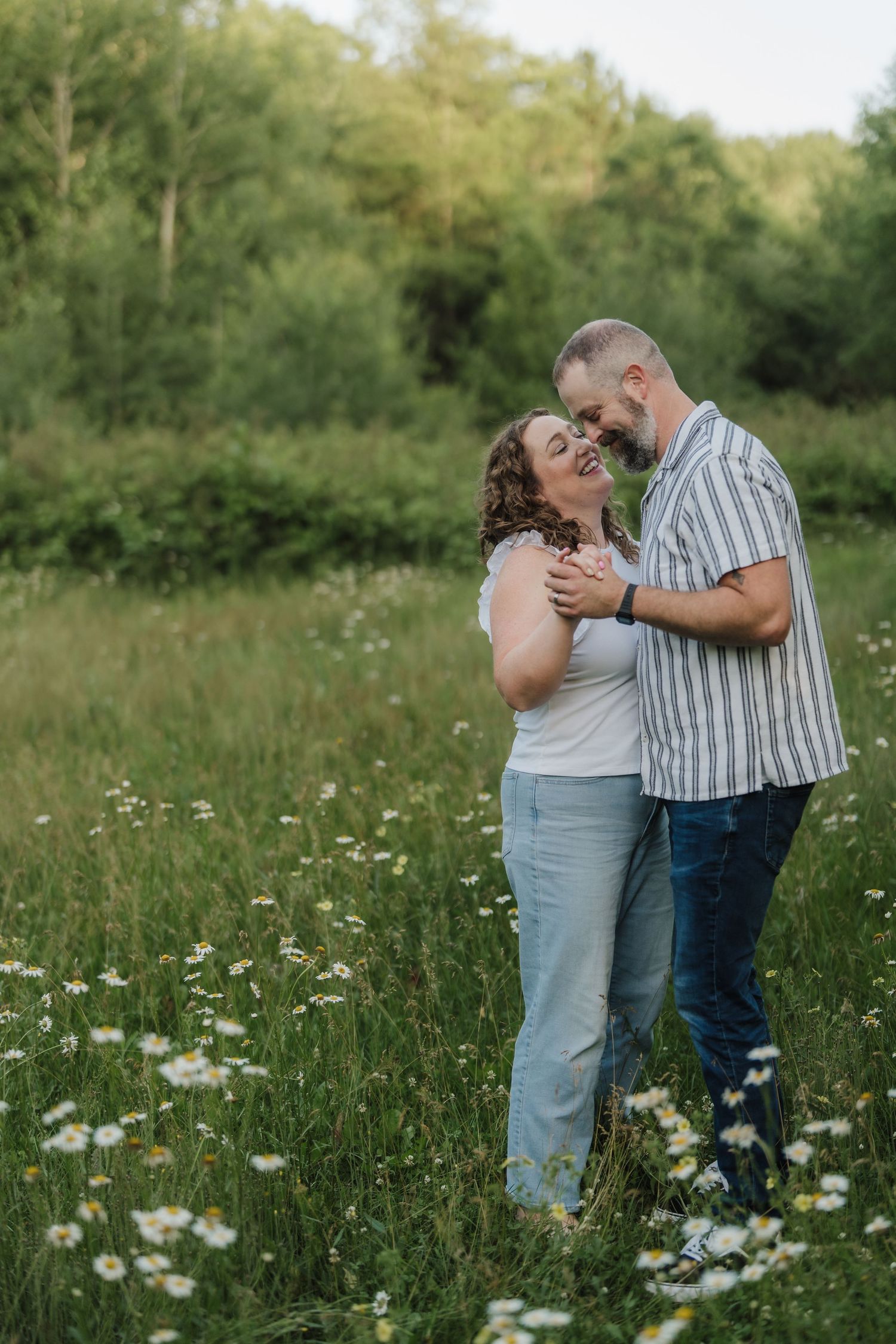 A smiling couple embraces tenderly in a wildflower meadow surrounded by lush green trees.