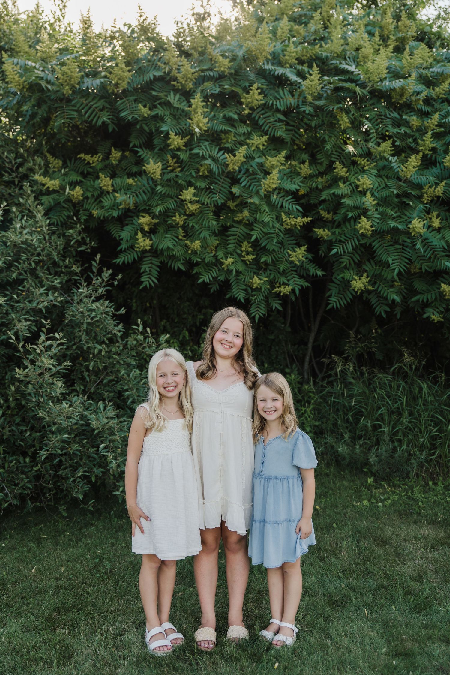 Three young girls in pastel dresses smiling together outdoors in front of lush green bushes.