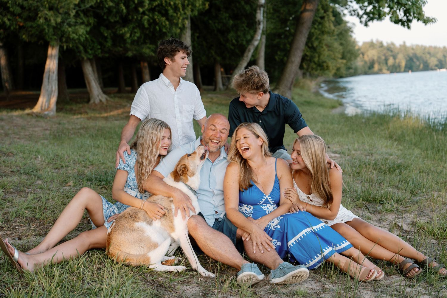 Happy family of five laughing outdoors by a lake with a golden dog on a sunny day.