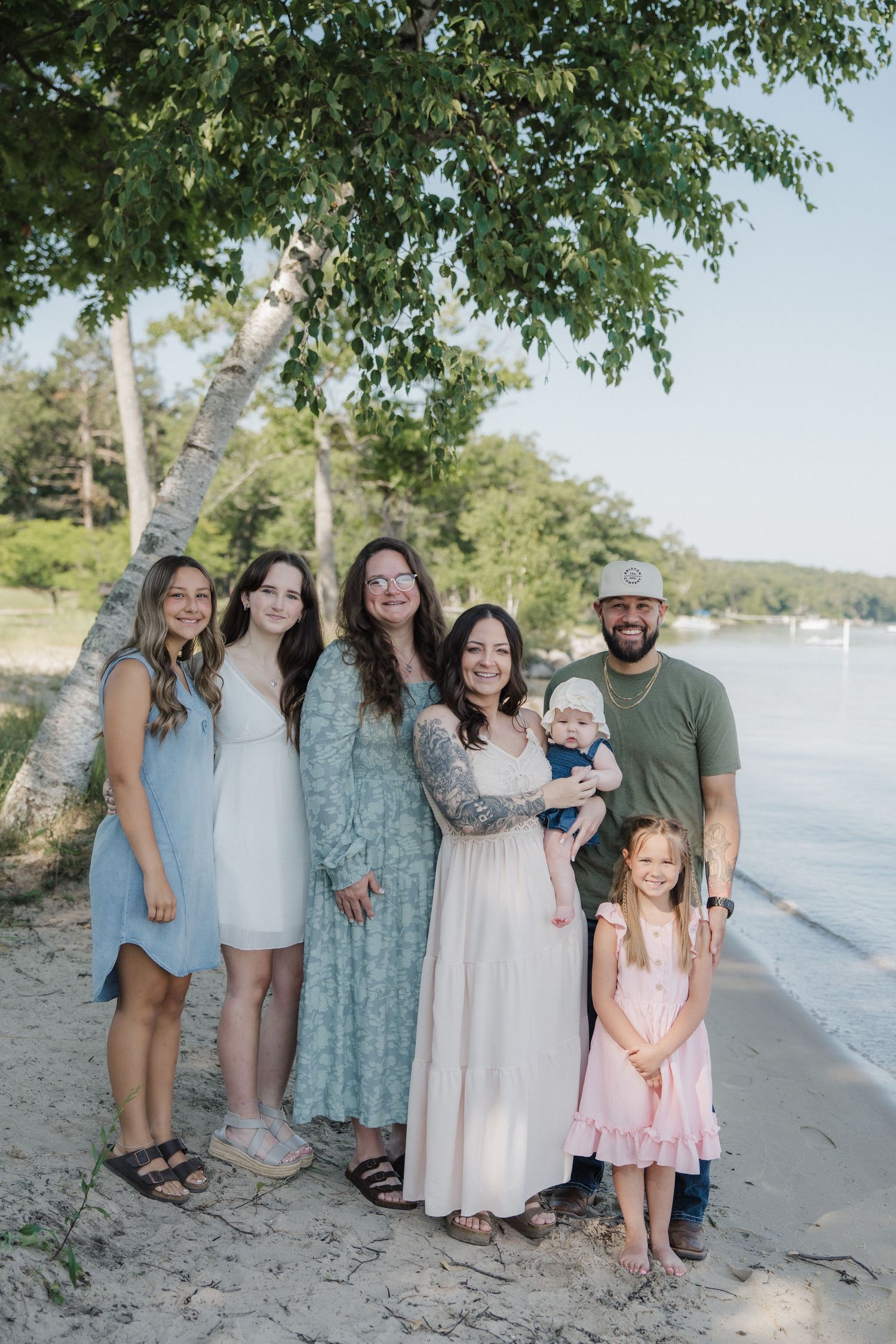 A joyful family of seven poses on a sandy beach near a lake, surrounded by lush green trees on a sunny day.