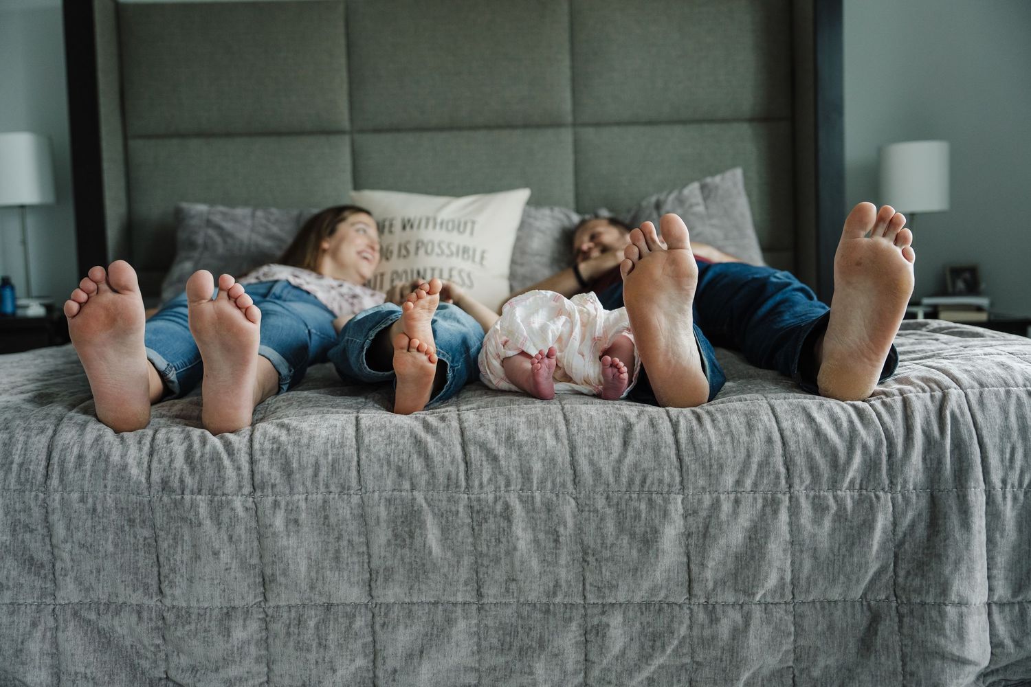 Family of three relaxing on a gray bed, parents and baby lying with feet toward camera in cozy bedroom.