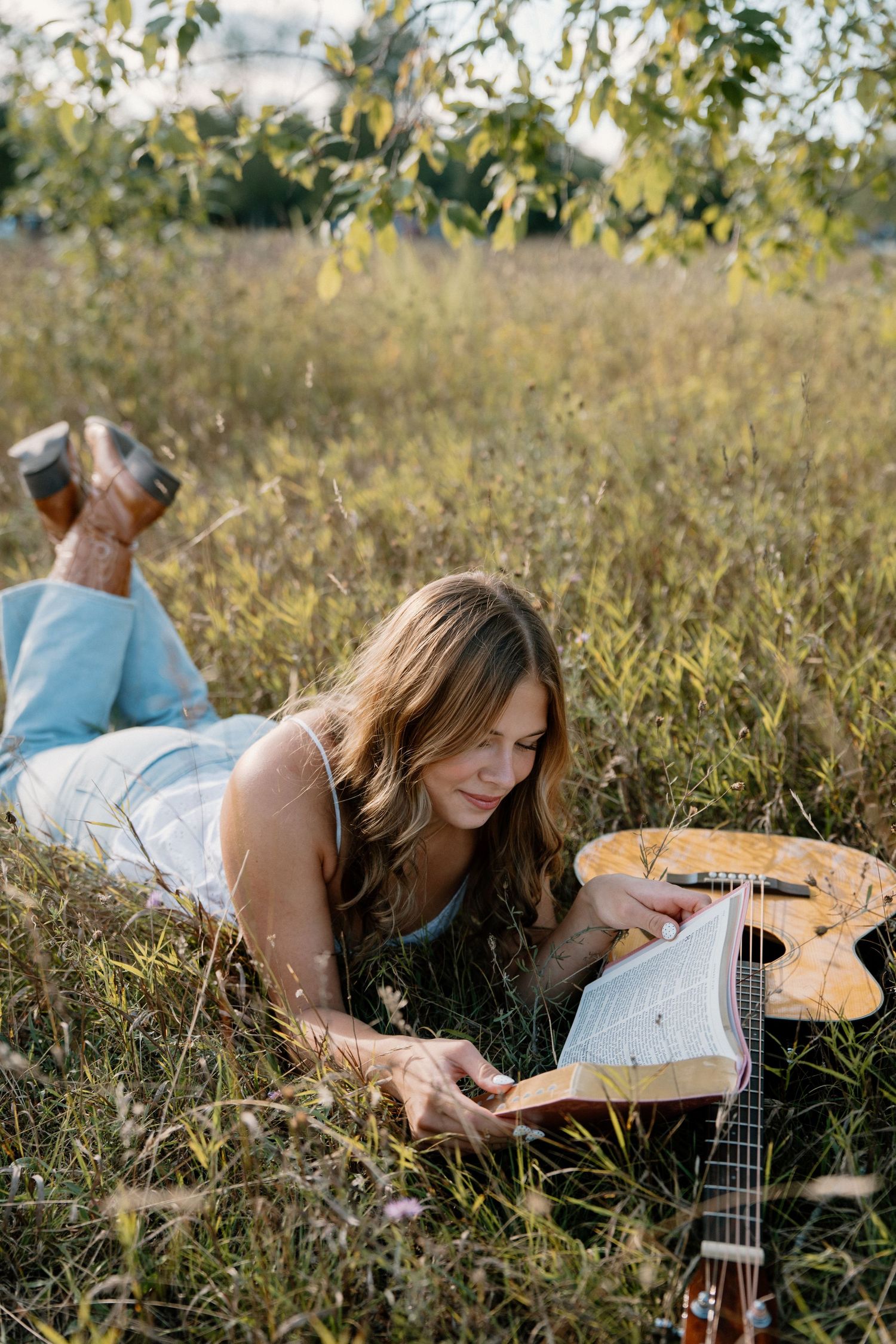Young woman lying in a grassy field reading a book beside an acoustic guitar on a sunny day.