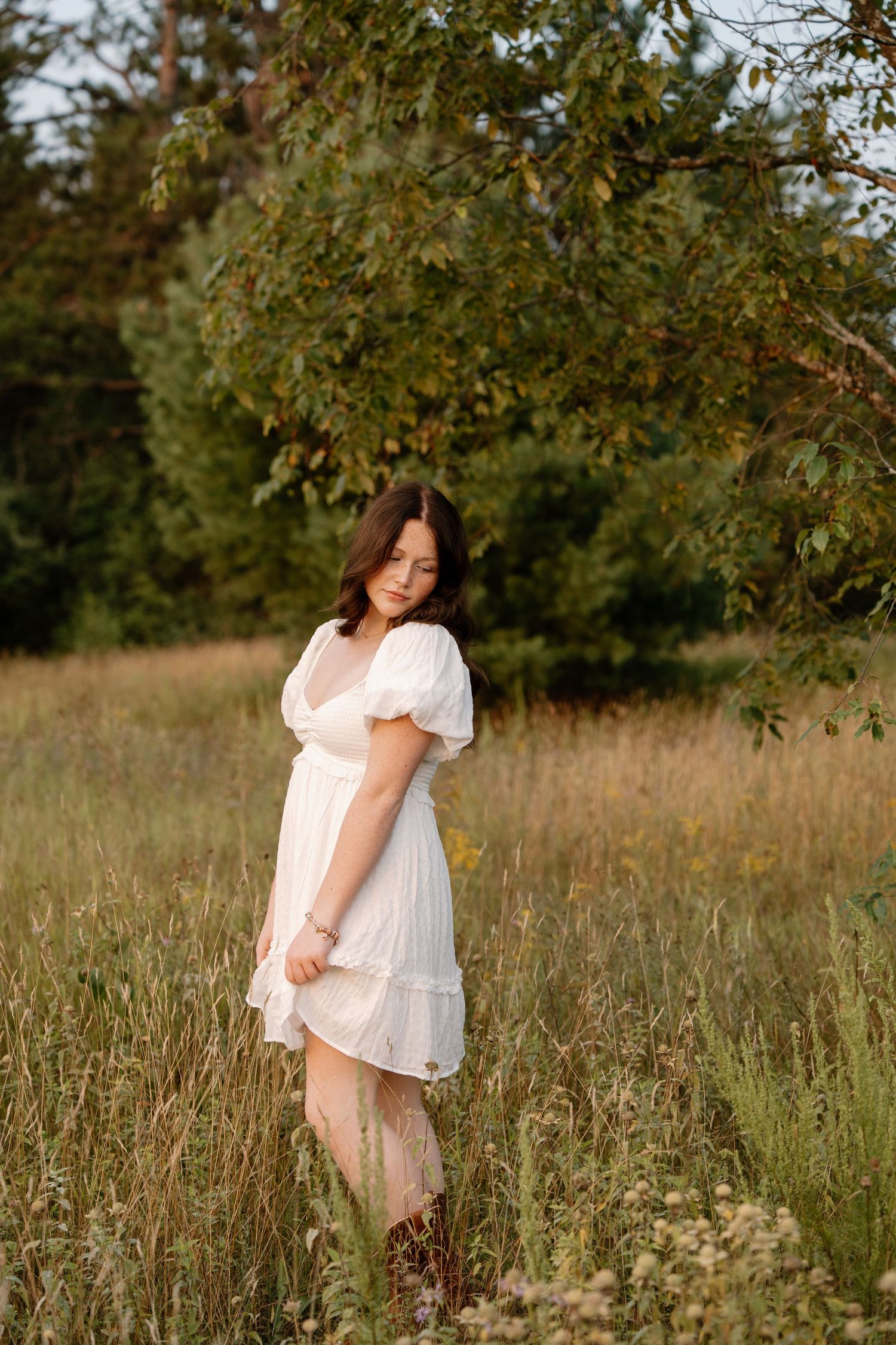 Young woman in white dress standing in golden field with green trees backdrop.