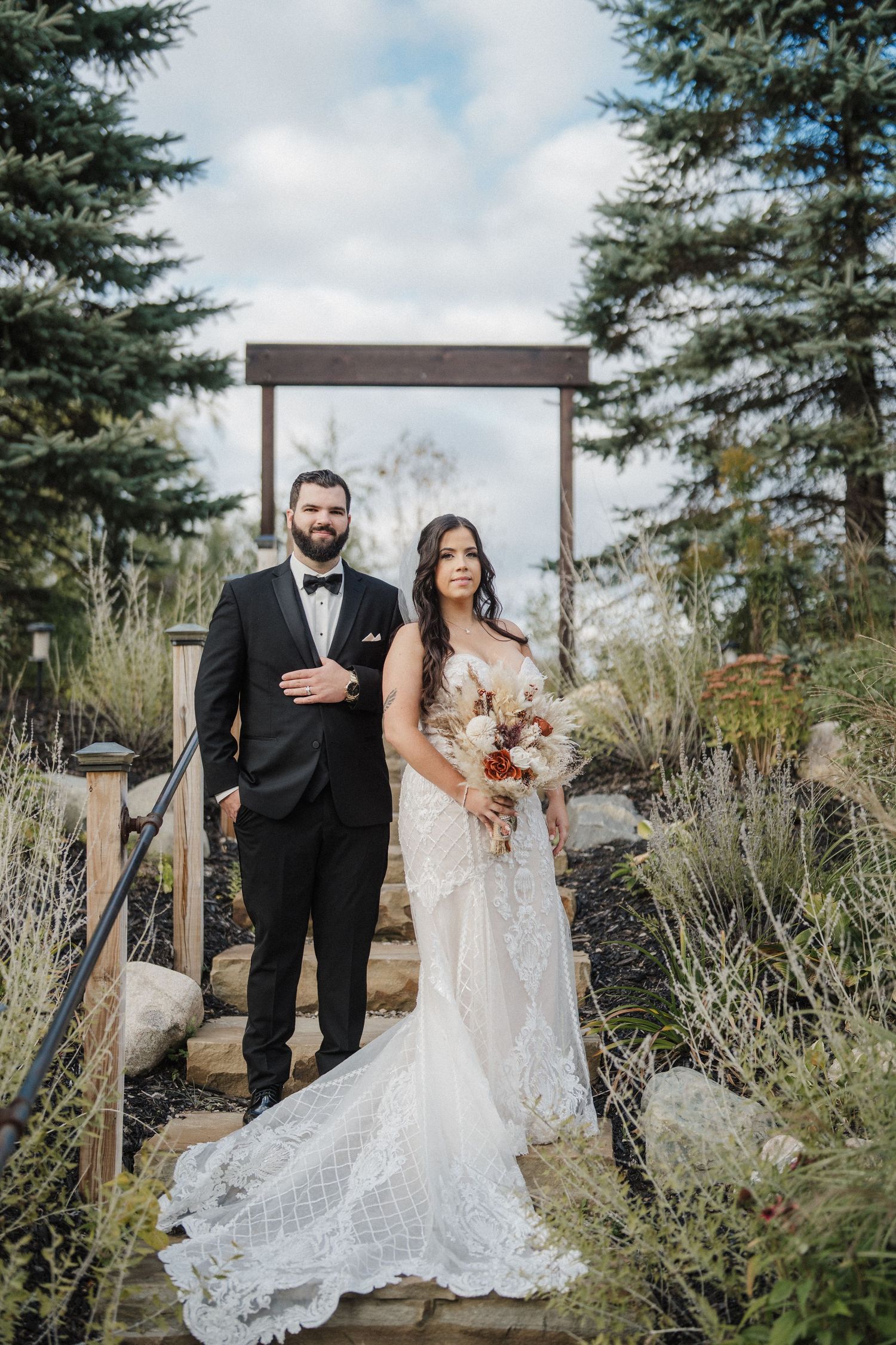 A wedding couple poses together in a garden setting with evergreen trees and ornamental grasses in the background.