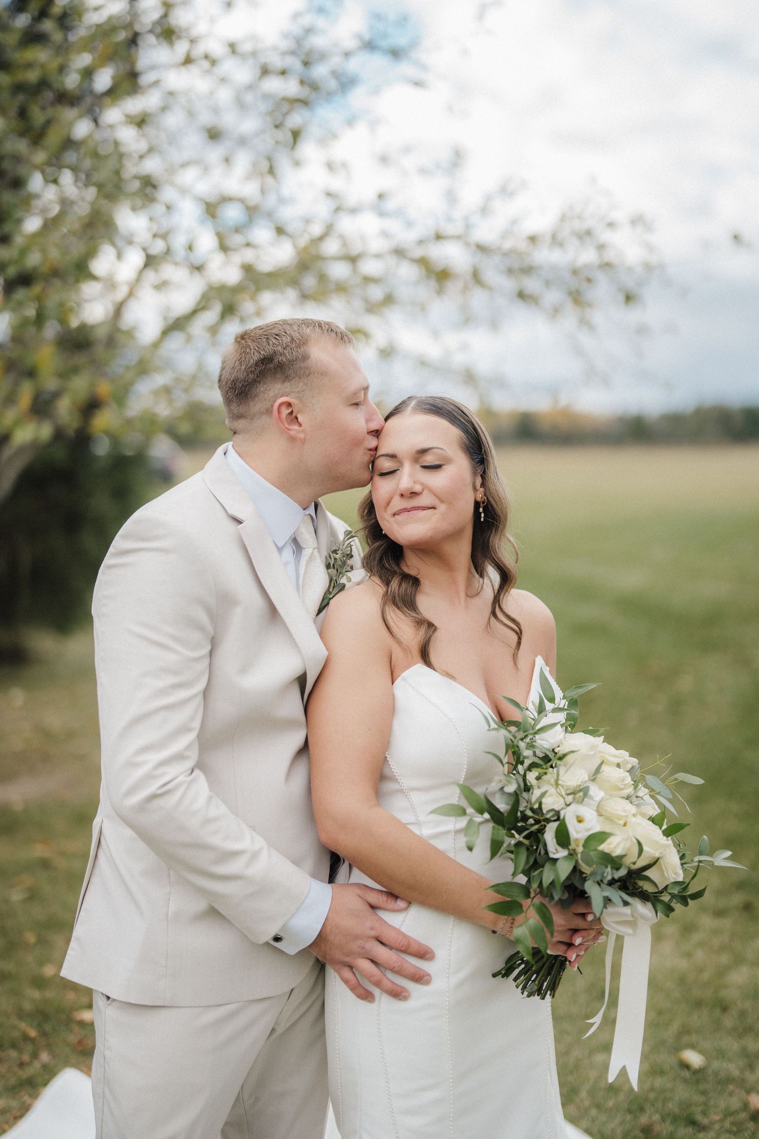 A romantic outdoor wedding photoshoot in a grassy field with white floral bouquet and light-colored attire.