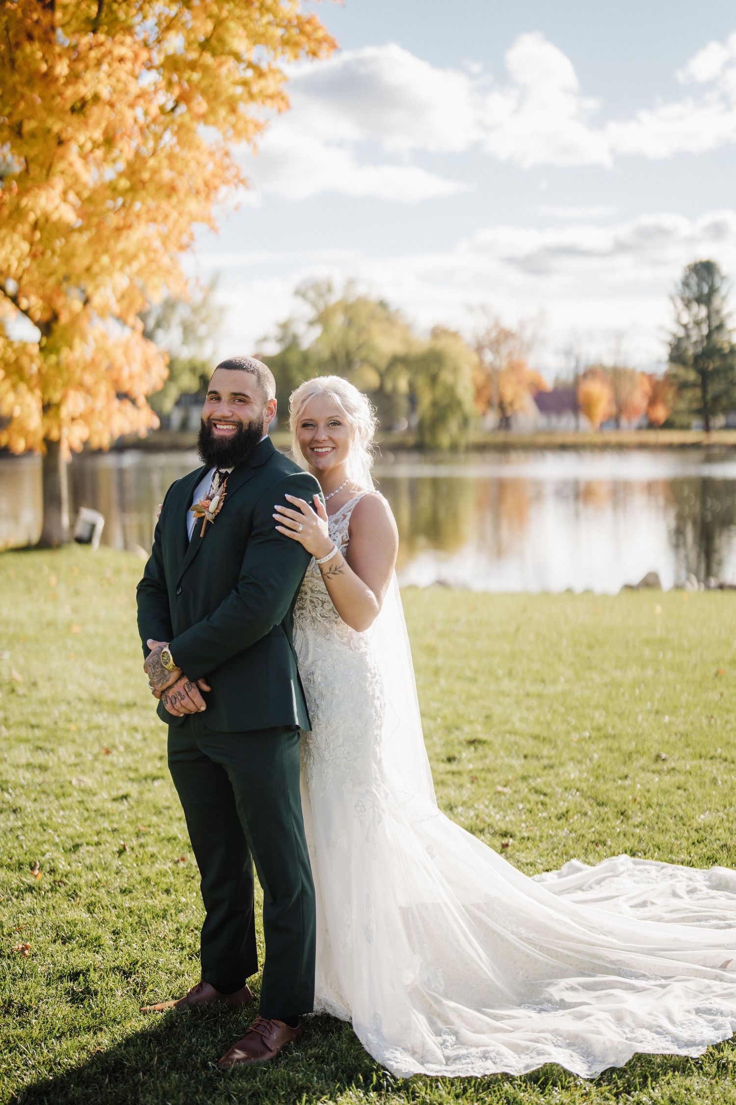Newlyweds share tender moments on a grassy lawn beside a lake during fall season.