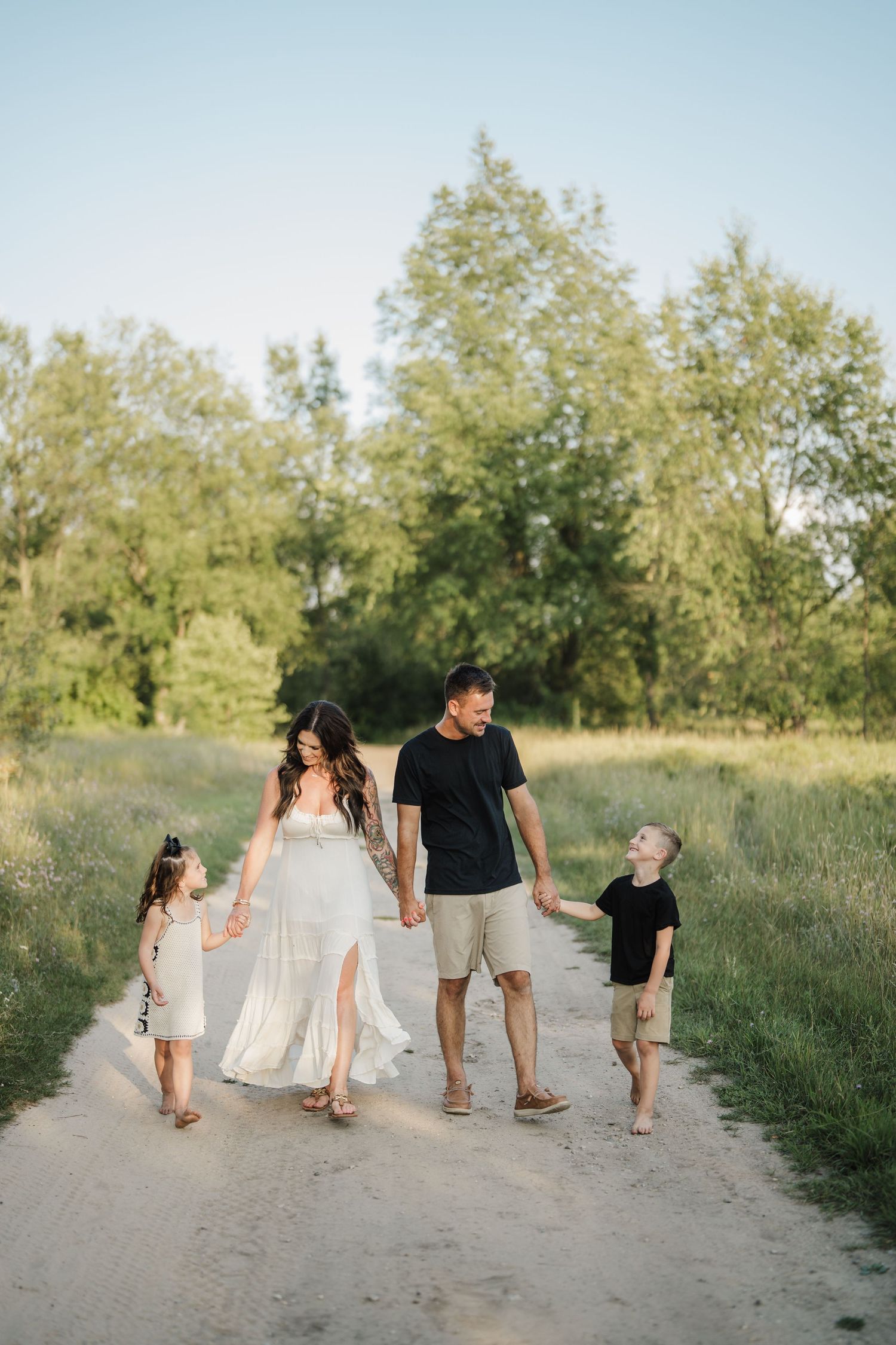 A family walks together holding hands on a dirt path surrounded by green grass and trees in warm sunlight.