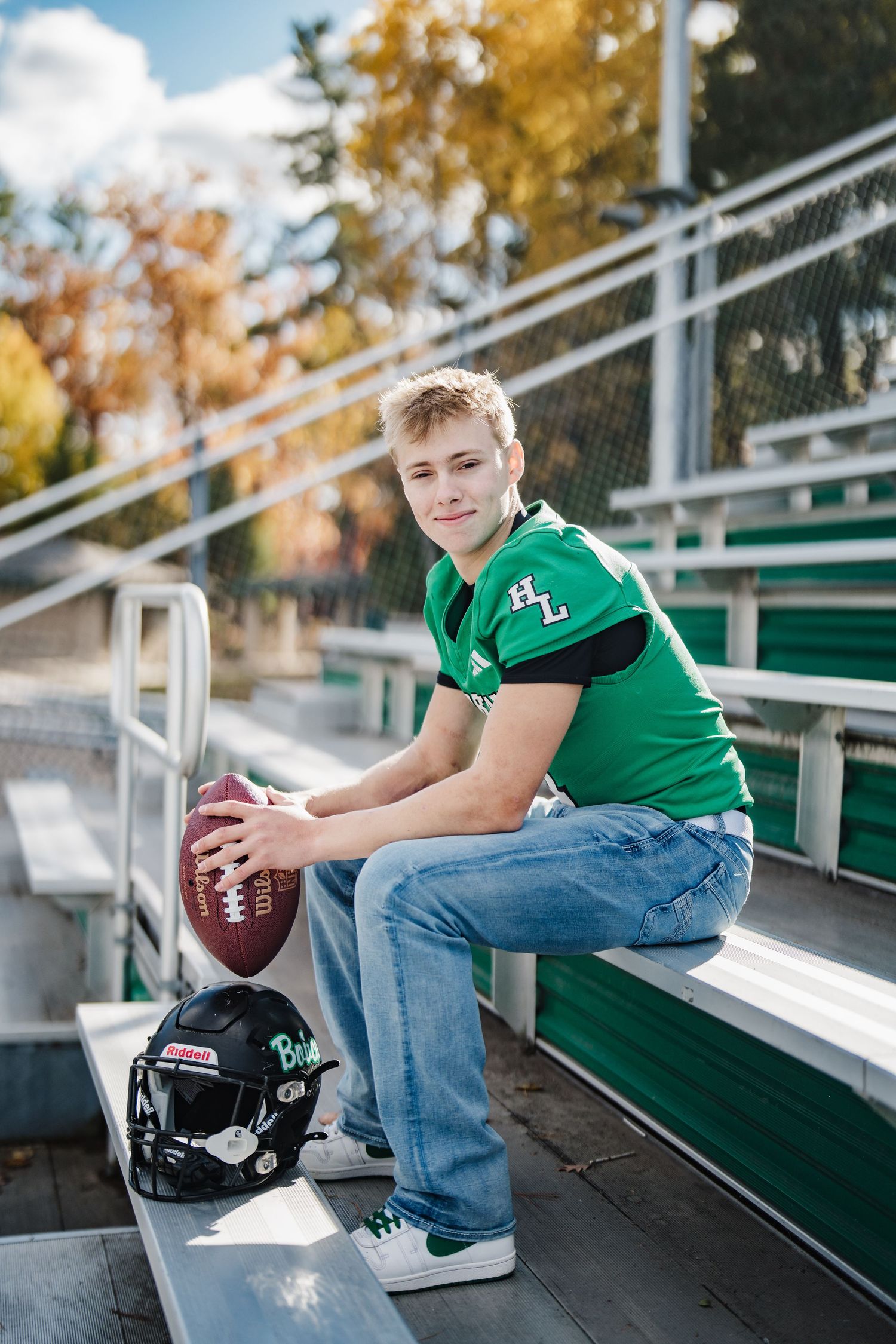 Football player in green jersey sits on stadium bleachers with helmet and ball during fall season.