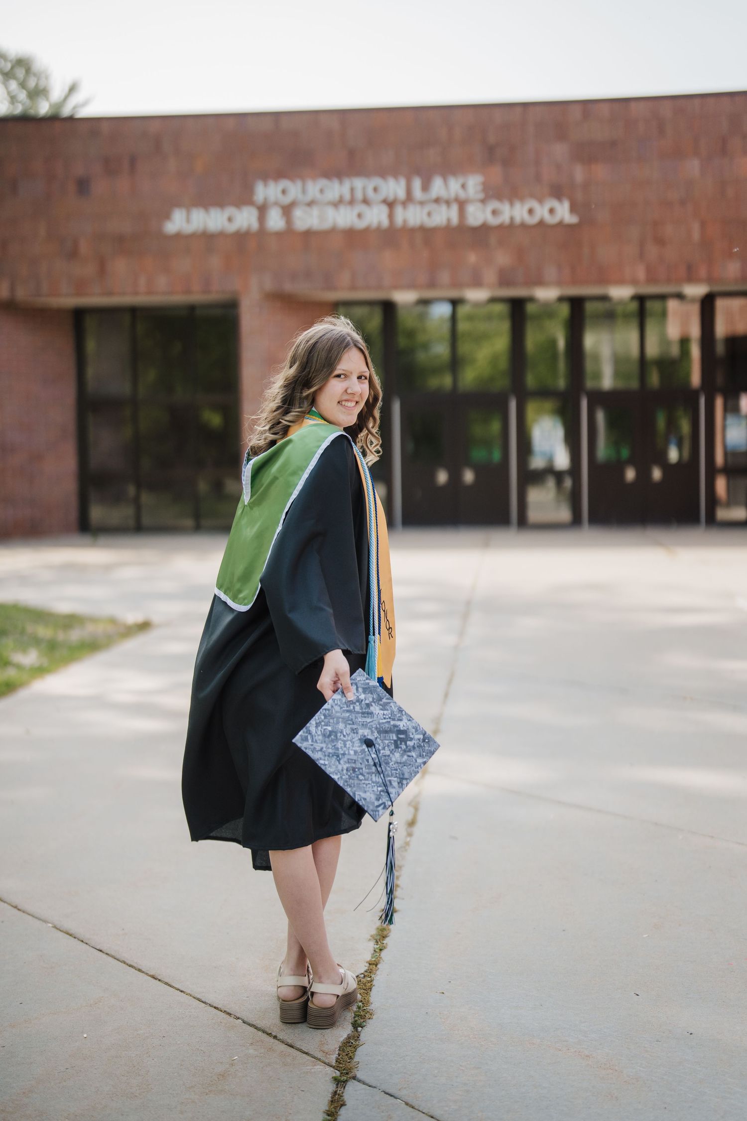 Graduate in black dress and cap holds diploma while posing in front of Houghton Lake Junior & Senior High School.