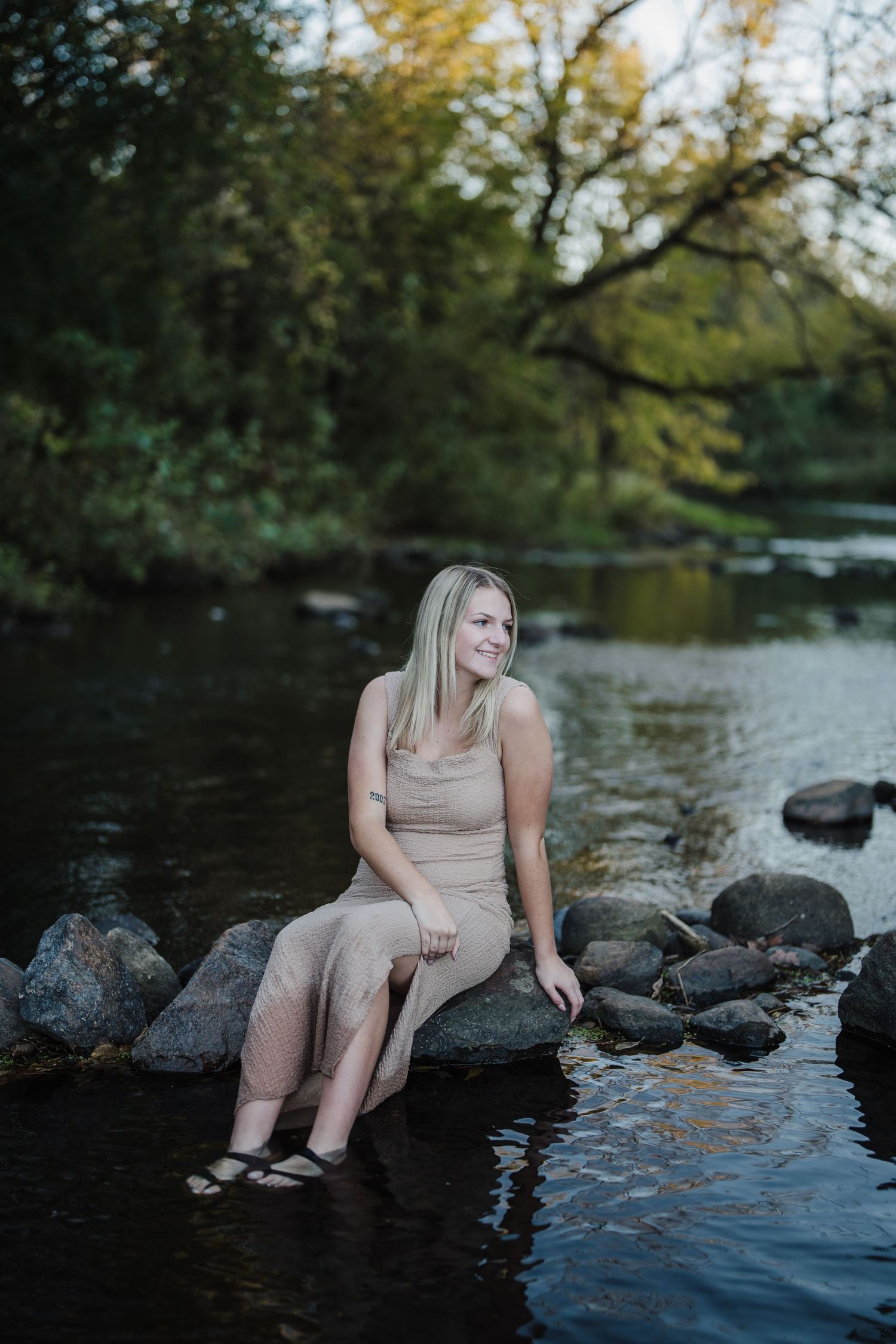 A figure sits on dark rocks by a tranquil lake at dusk wearing a beige outfit as trees reflect in the calm water.