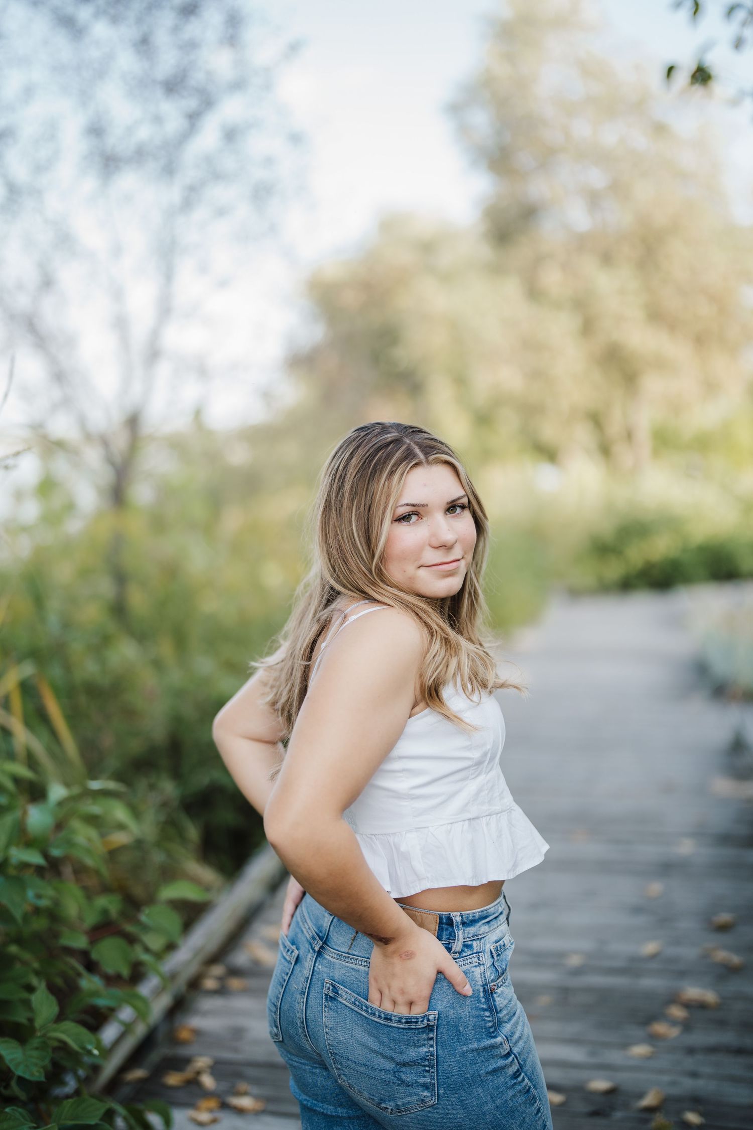 A person in a white crop top and blue jeans poses outdoors on a path surrounded by greenery.