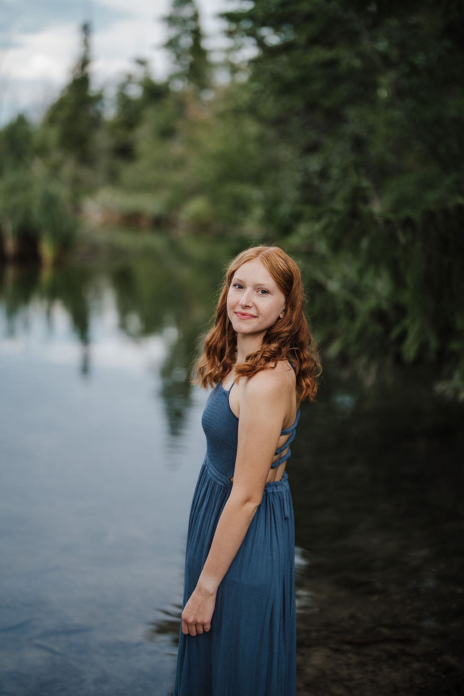 A serene lakeside portrait features someone in a flowing navy dress against a backdrop of evergreen trees.