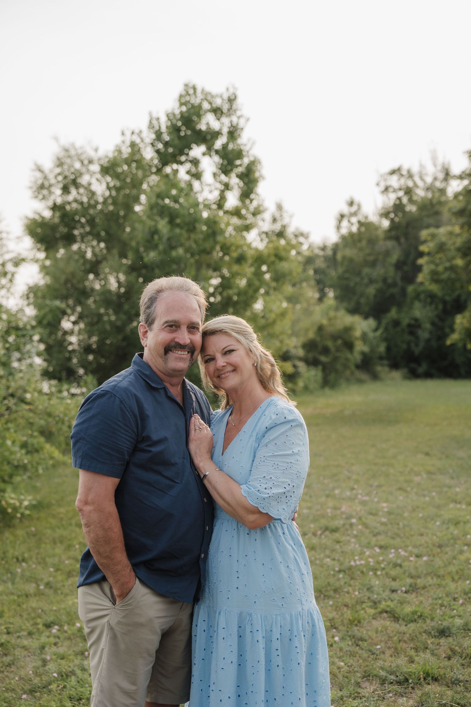 A smiling couple poses outdoors on a green lawn, woman in light blue dress leaning on man in navy shirt.