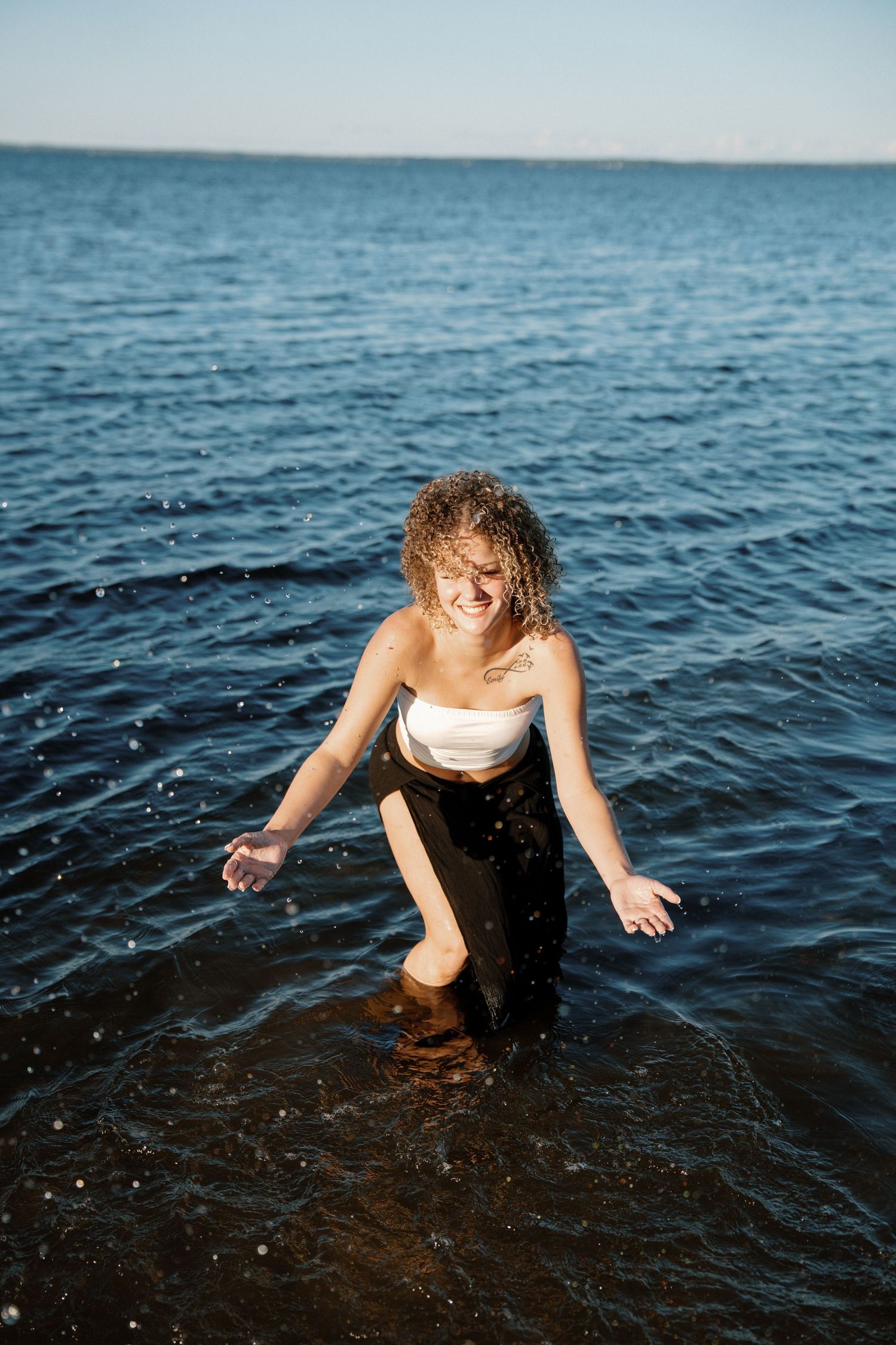 A smiling young woman with wet curly hair stands in the ocean wearing a white top and black pants.