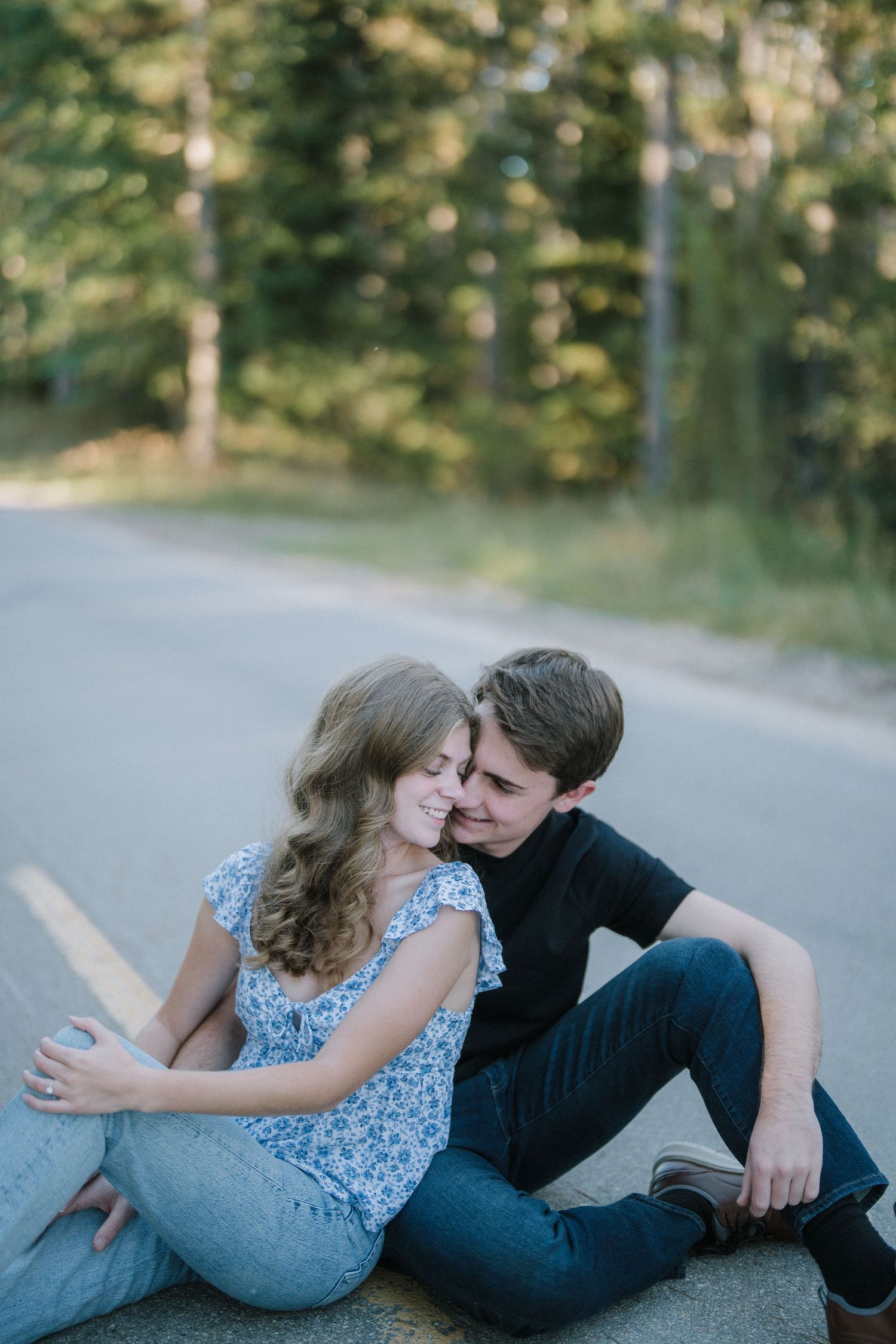 A couple sits intimately together on a rural road surrounded by trees in soft dreamy lighting.