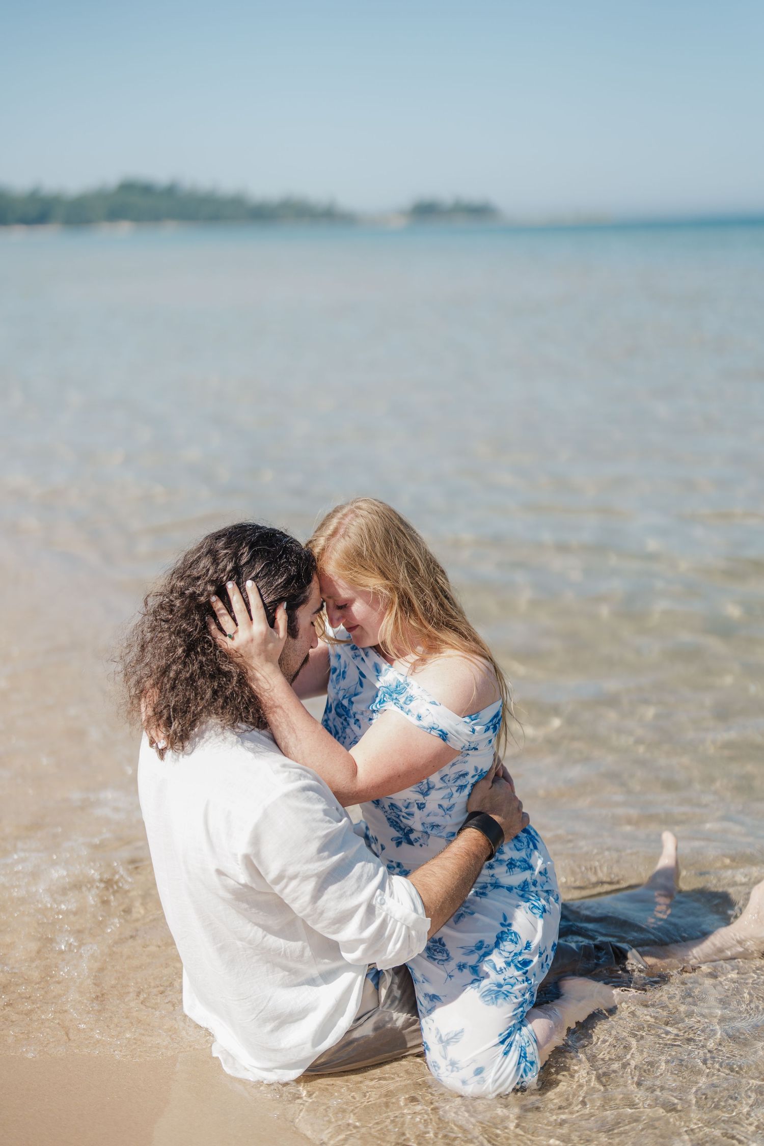 Two people in white and blue outfits sit close together on a sunny beach with crystal clear water in the background.