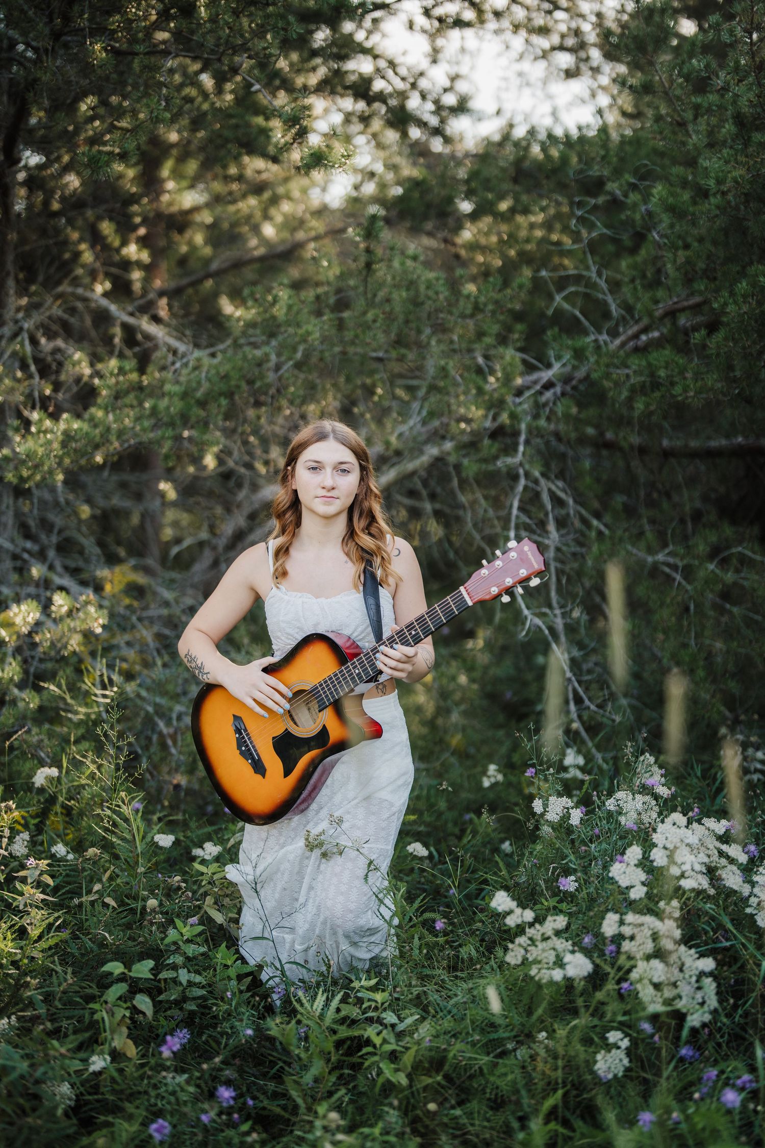 A person in a flowing dress plays acoustic guitar while sitting in a wildflower meadow at sunset.
