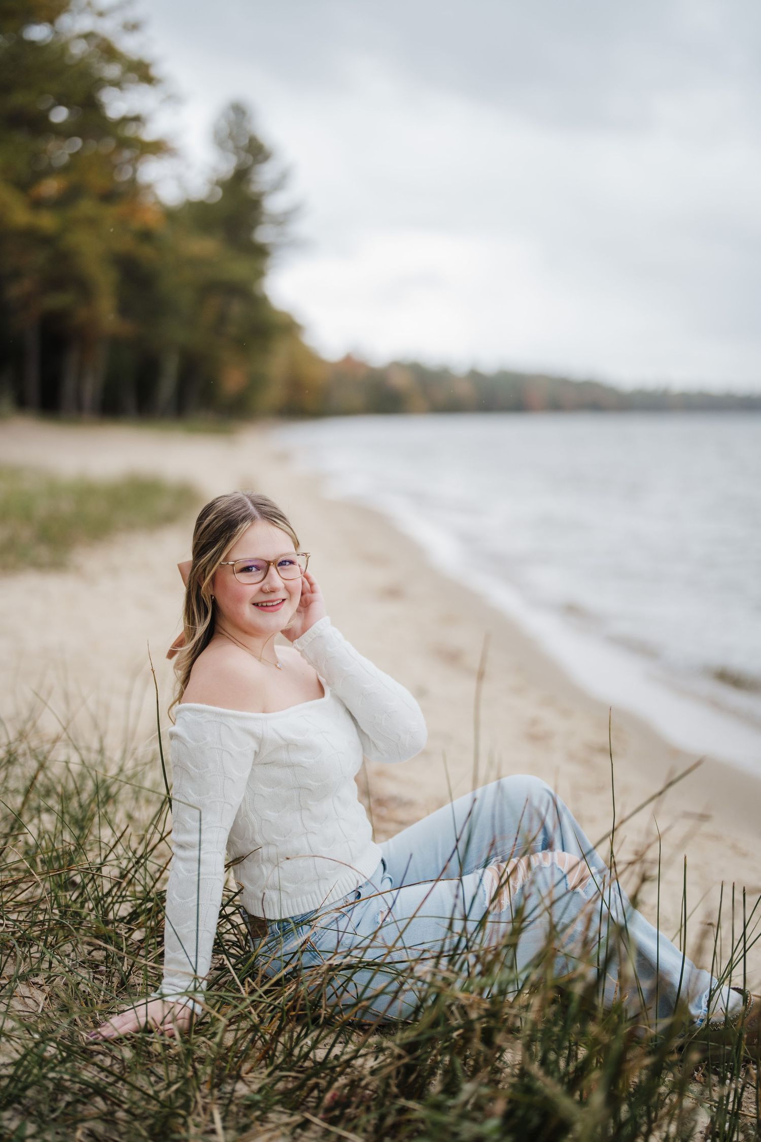 Beach portrait photography series showing someone in a white sweater and jeans reclining on sandy shore with beach grass.