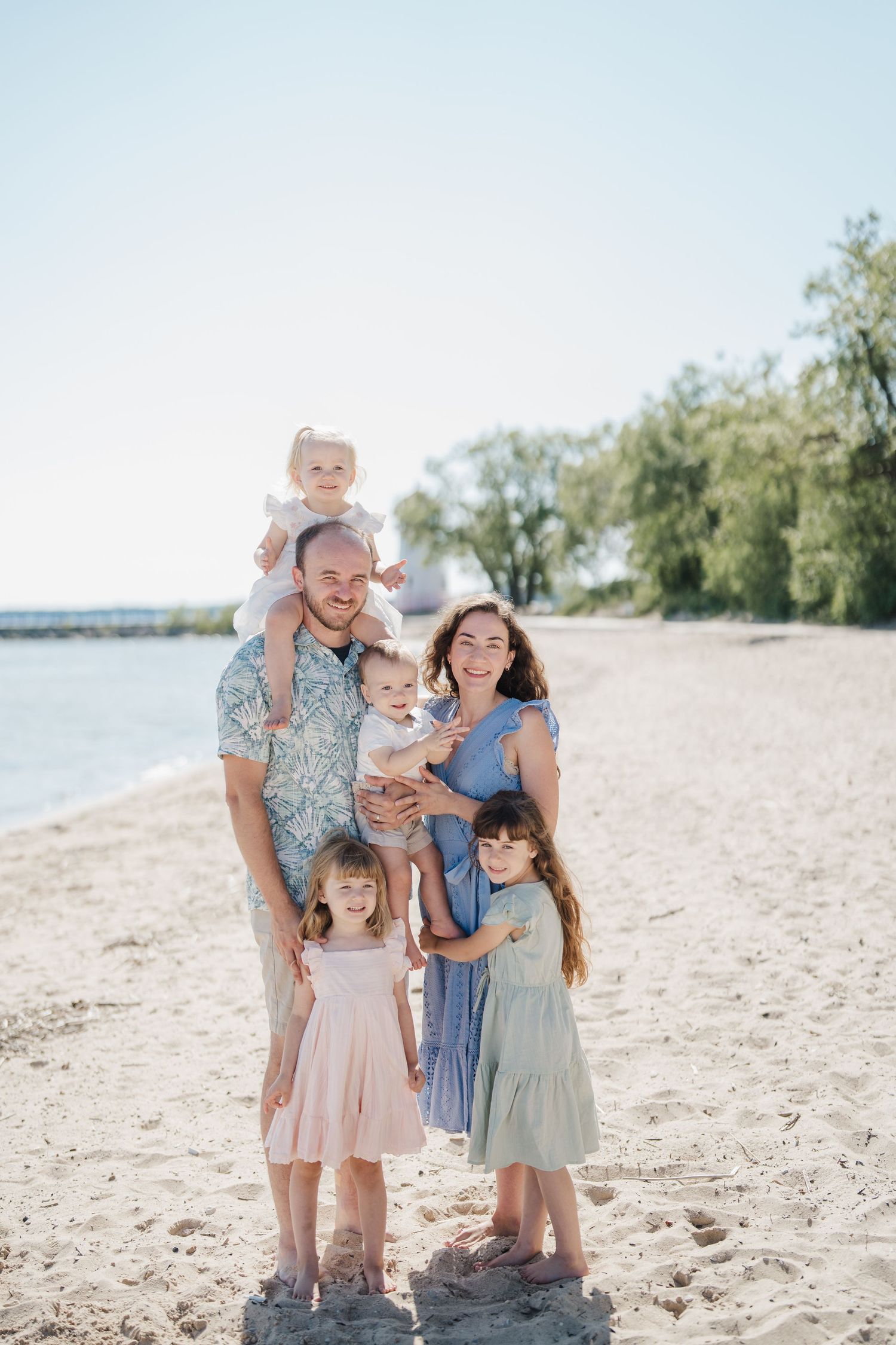 Family wearing coordinated pastel outfits poses together on a sunny beach with water and trees in background.