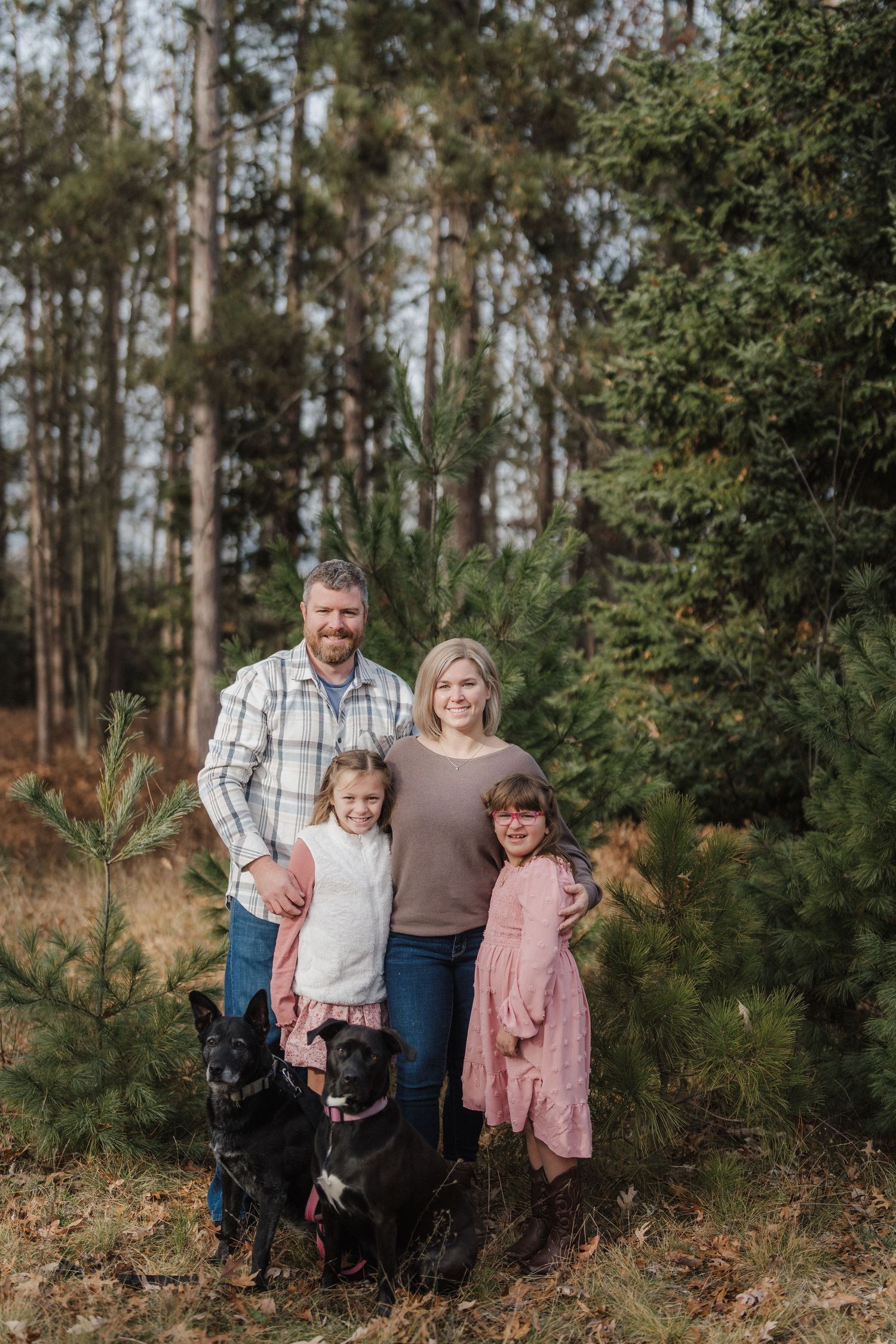 A family in coordinated casual outfits poses together in a wooded outdoor setting with pine trees in the background.