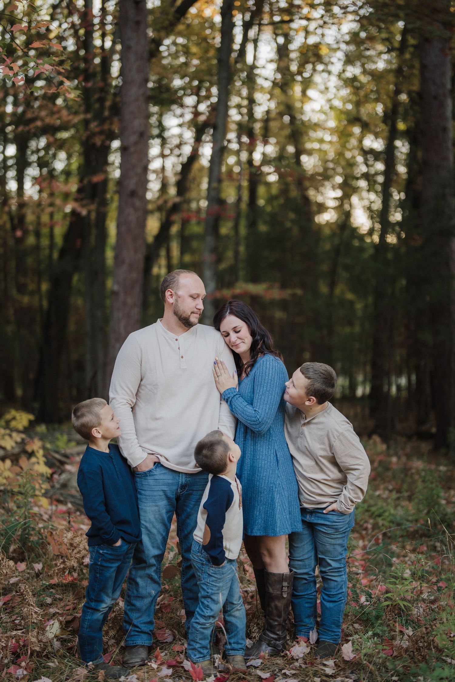 A family in coordinating blue and beige outfits poses together in a wooded forest setting during autumn.