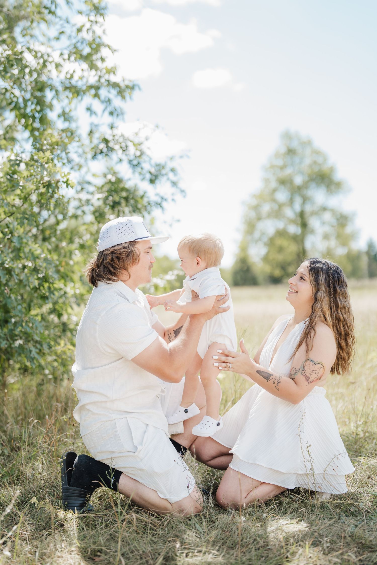 Family wearing white outfits sits together in a sunny meadow with birch trees on a bright summer day.