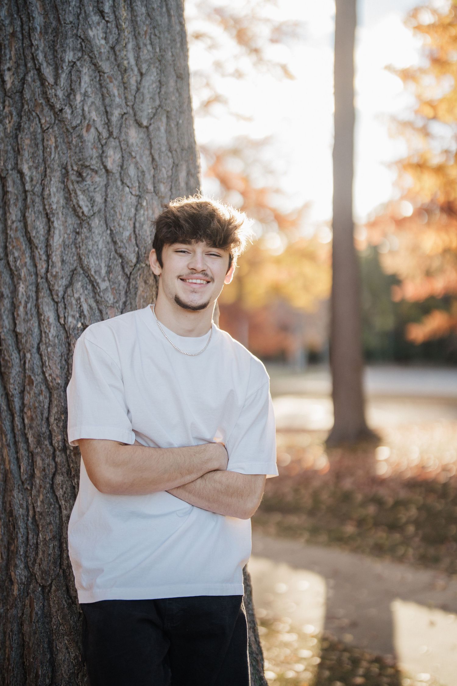 Portrait of someone in a white t-shirt leaning against a large tree trunk in autumn lighting.