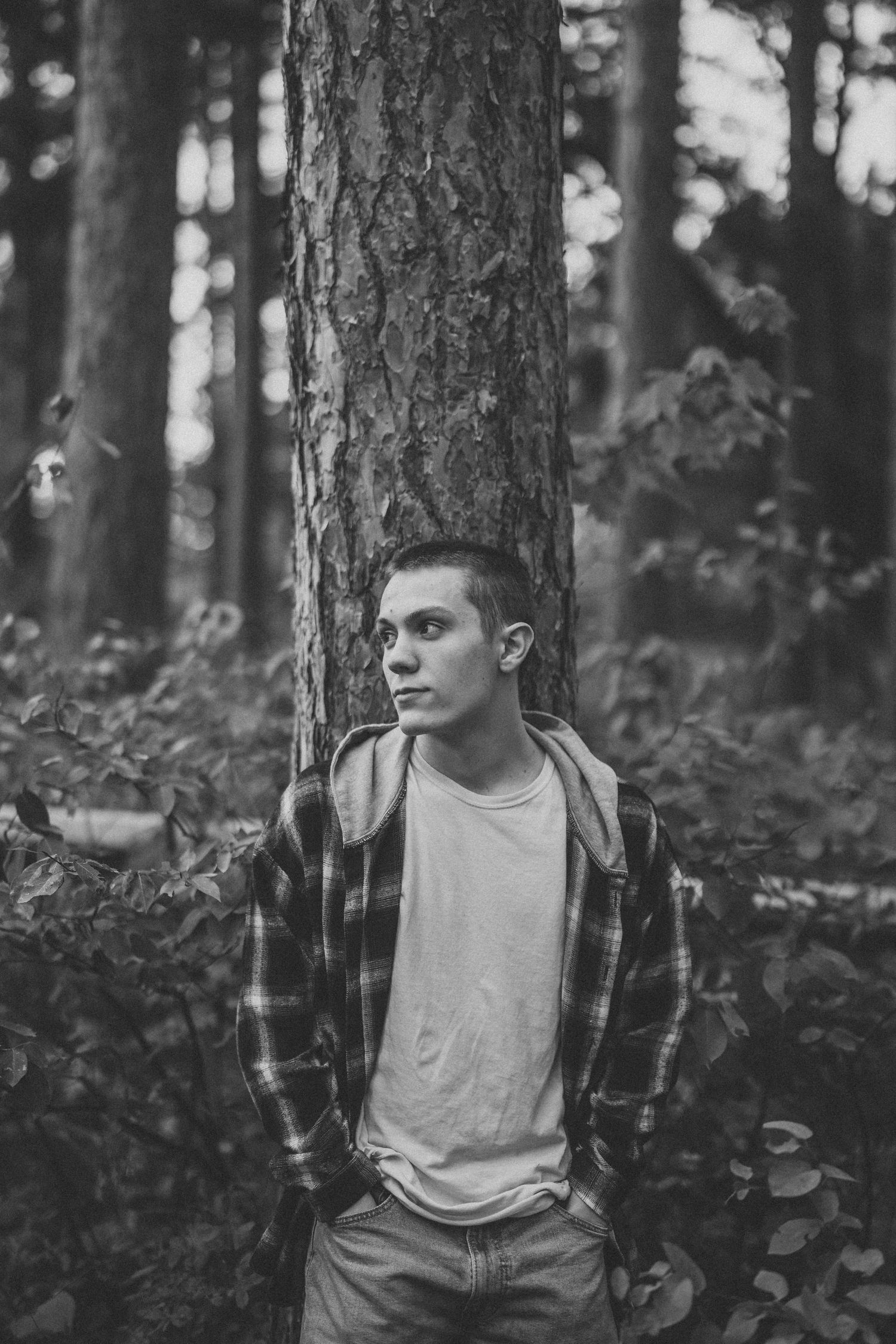 Black and white portrait of someone in a plaid flannel shirt and backpack standing against a tree trunk in a forest.