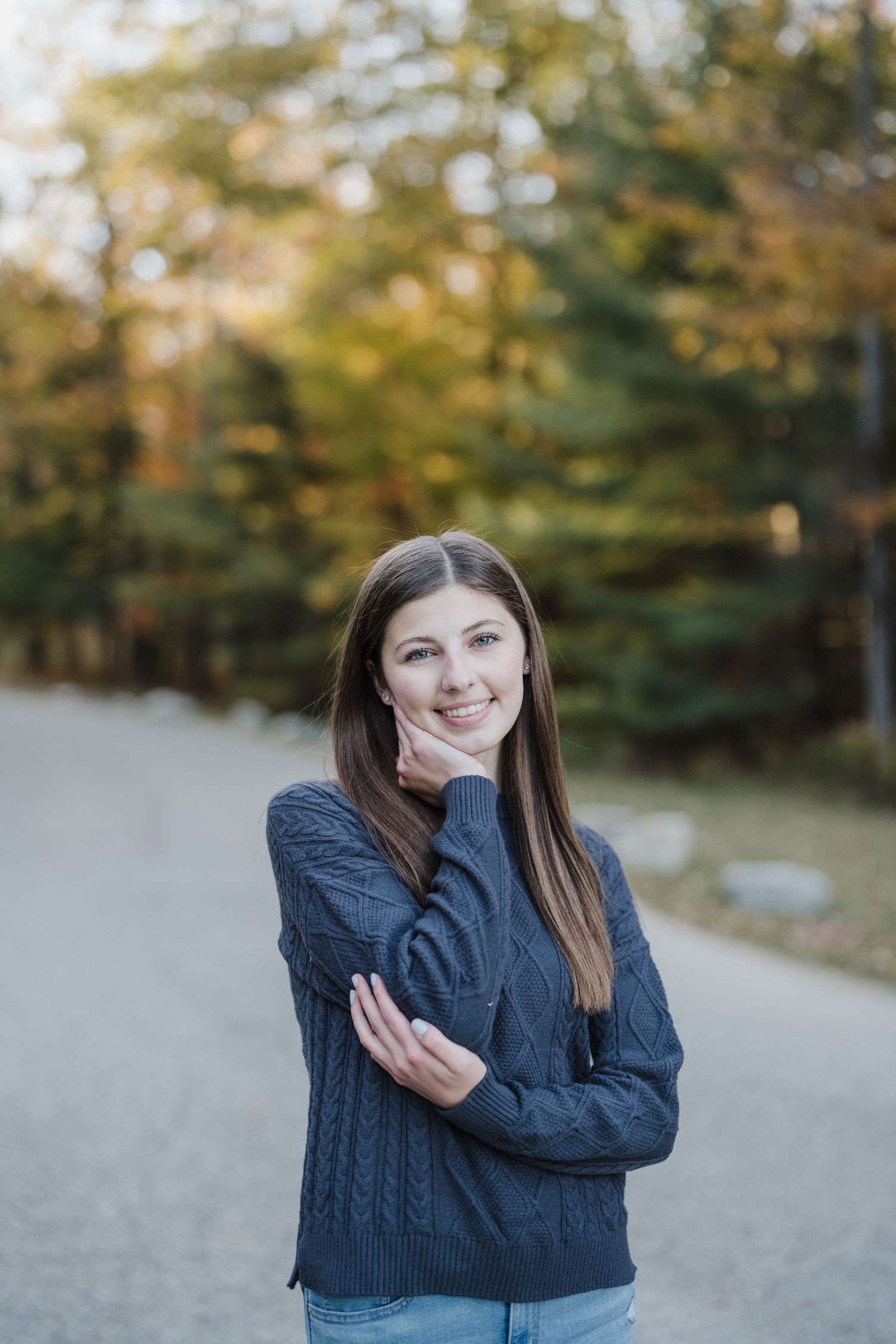 A person in a dark sweater poses outdoors against a blurred autumn background on a path.
