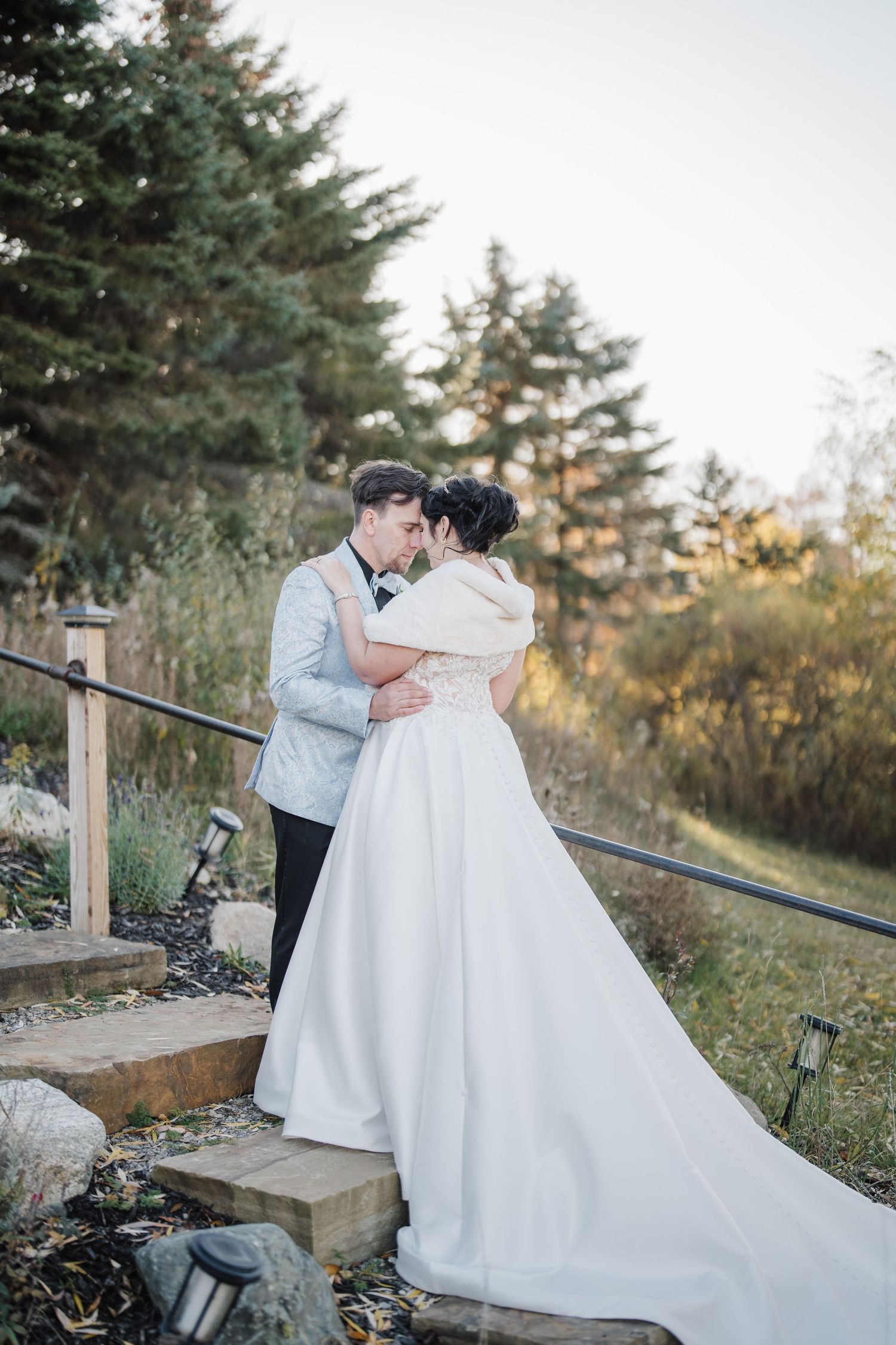 A romantic wedding couple embraces on rustic wooden steps surrounded by evergreen trees at sunset.