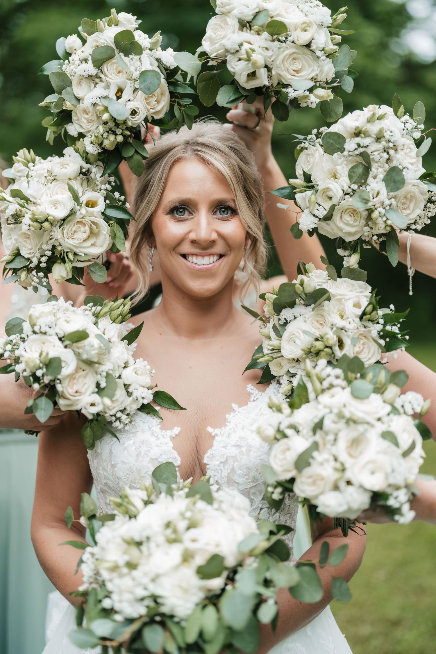 Bride smiles surrounded by elegant white floral bouquets with greenery outdoors.
