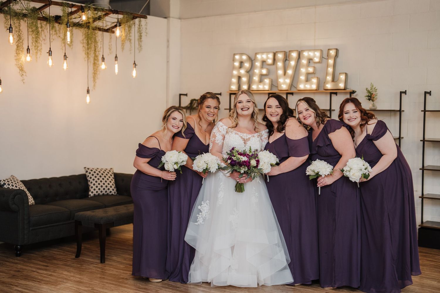 Wedding party in purple bridesmaid dresses poses together under illuminated Revel sign.